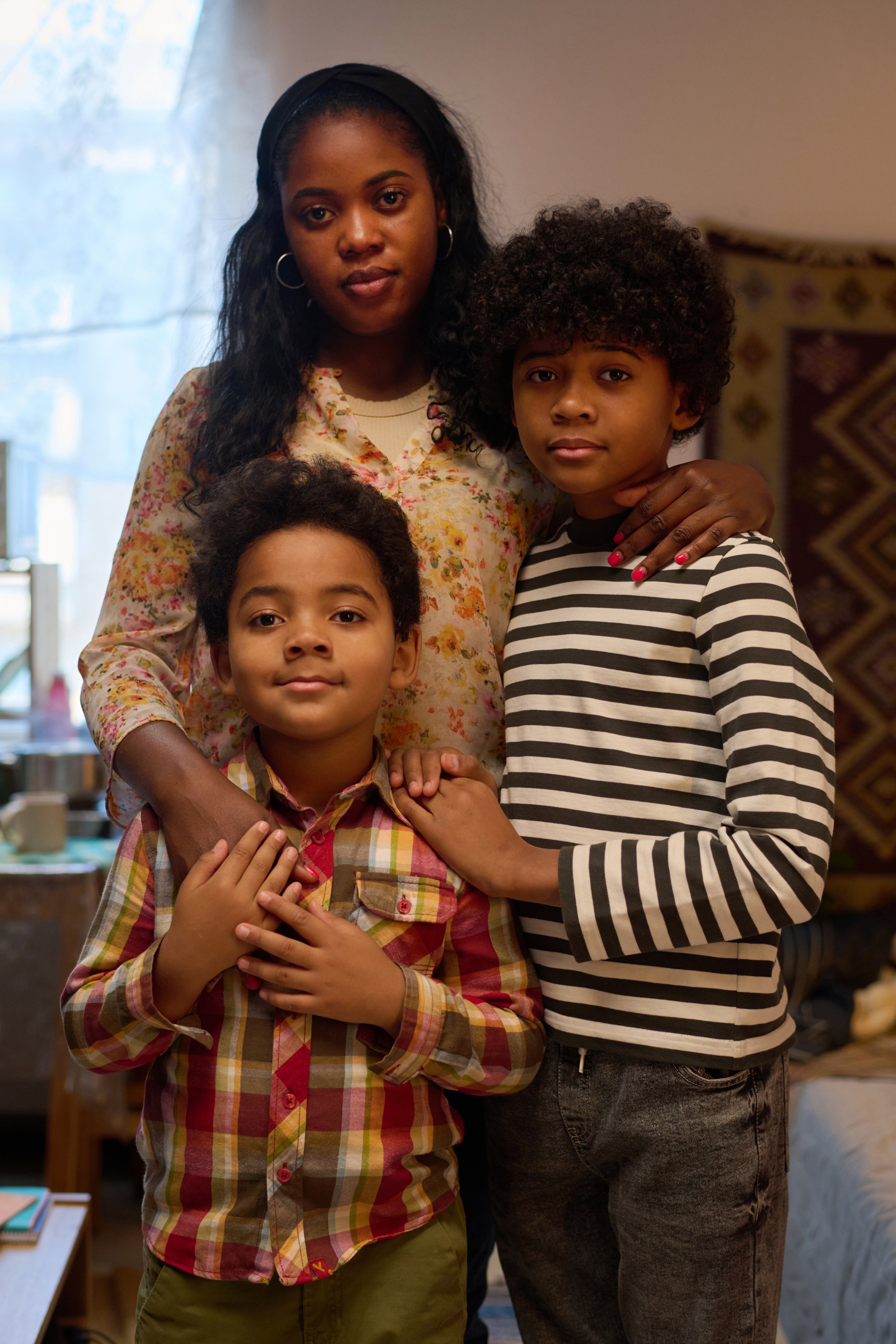 A woman with two children in a cozy room, all looking at the camera. The woman has long dark hair and is wearing a floral blouse. The boy in front has curly hair and is wearing a plaid shirt, with his hands on his chest. The girl on the right has shorter curly hair and is wearing a black-and-white striped shirt, resting her hand on the boy's shoulder.