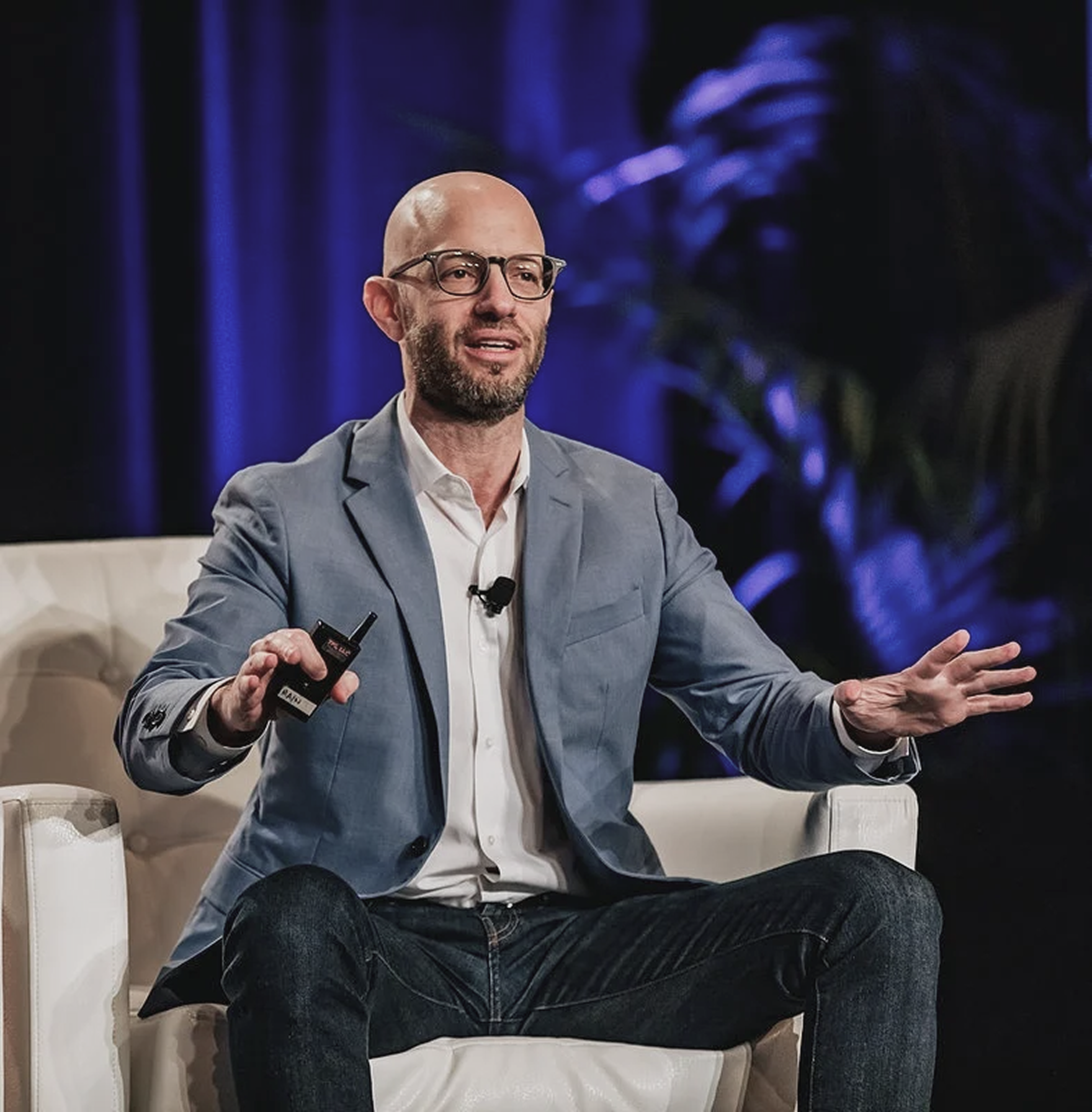 A man with glasses and a beard, wearing a light gray blazer and white shirt, is seated on a white chair on stage, speaking and gesturing with his right hand, while holding a small microphone in his left hand.