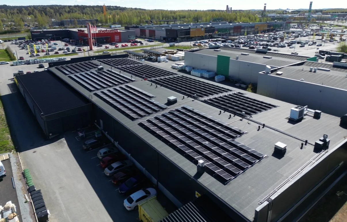 Rakennuksen katolla on rivistöjä aurinkokennoja, ja sen ympärillä on pysäköintiä. Kaupan ja ostoskeskuksen sisäänkäynneissä on liikennettä, ja taustalla näkyy paljon muita rakennuksia ja puita.