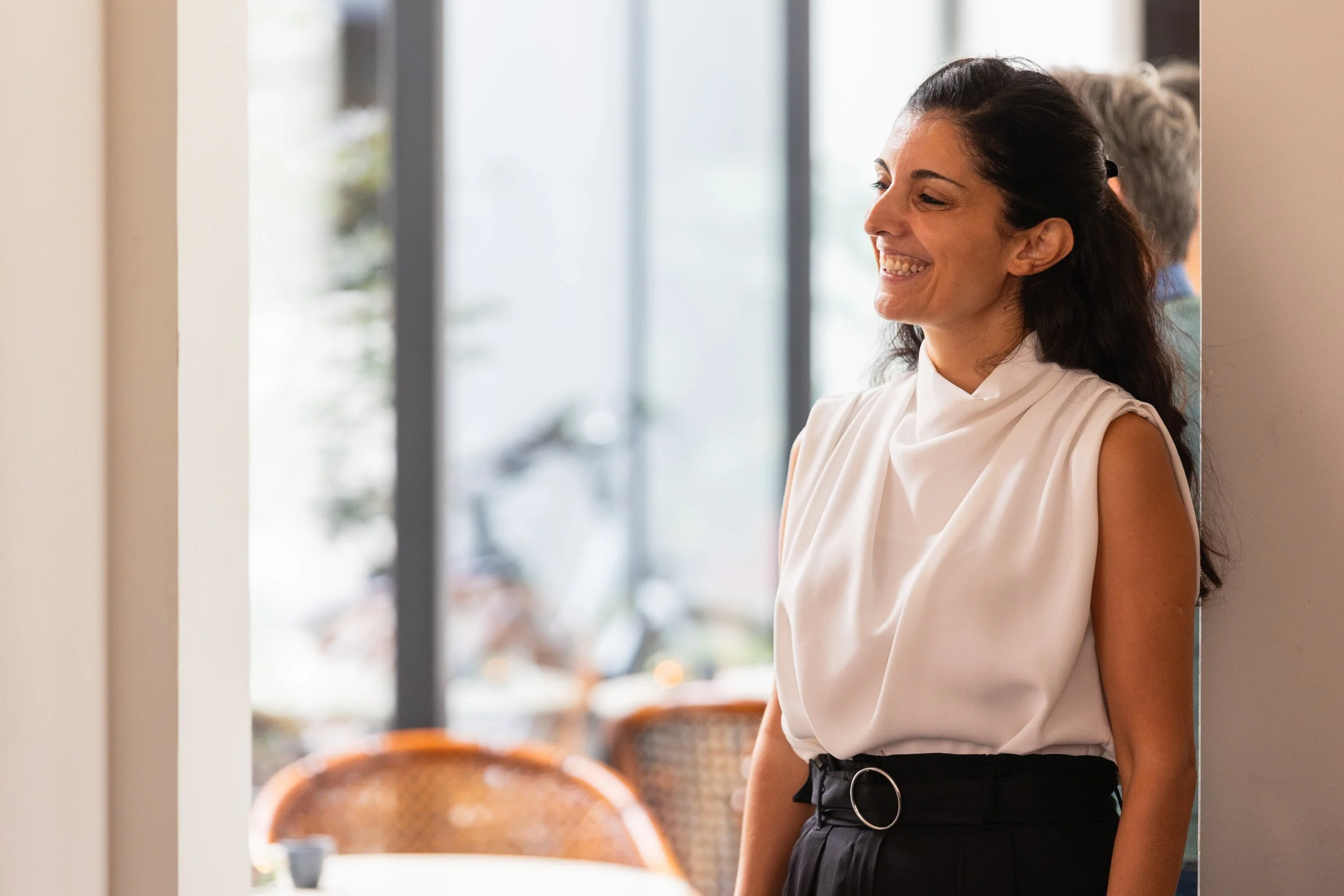 A woman with dark hair smiling, wearing a sleeveless white top and black high-waisted pants, standing in a bright room with large windows.