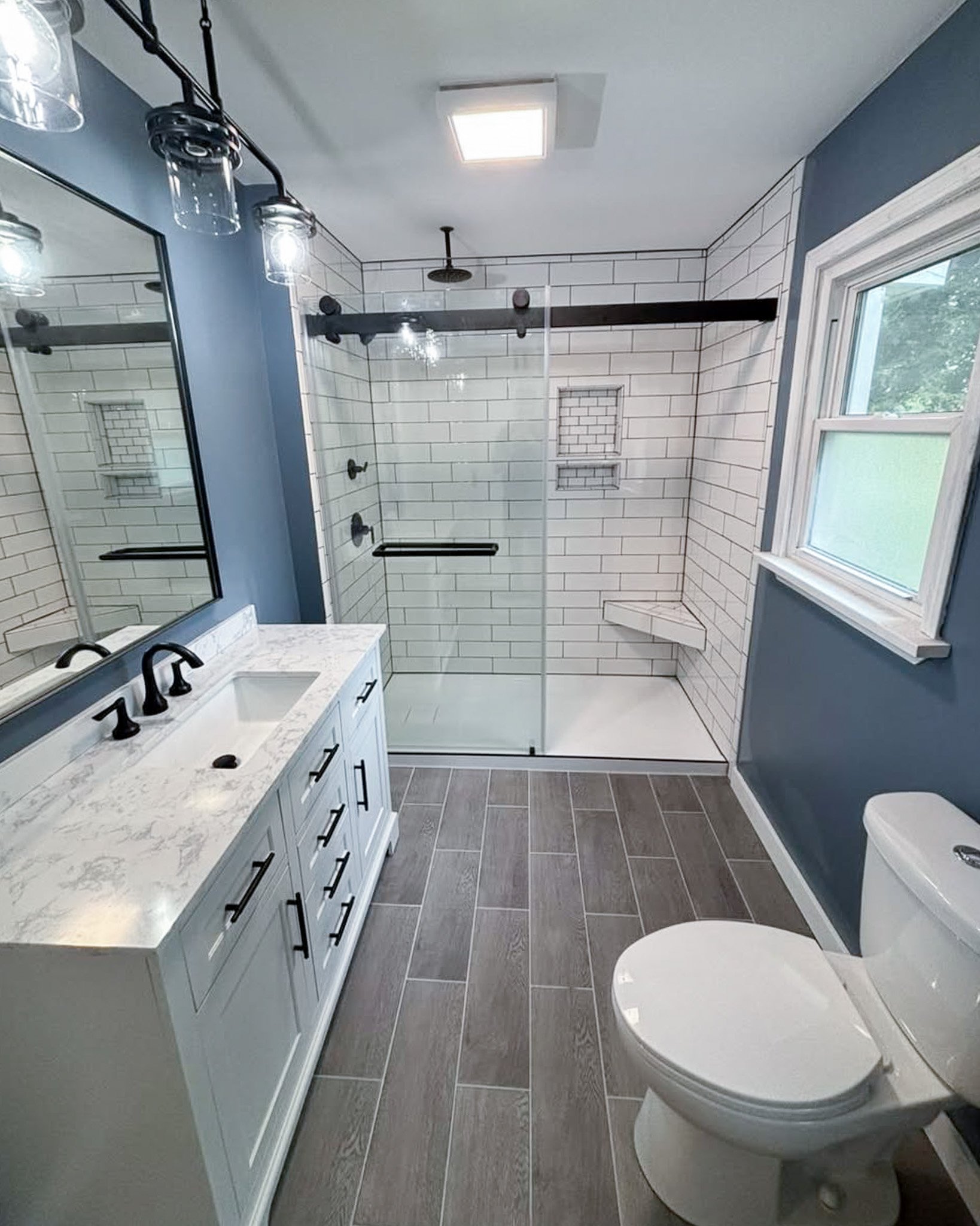Modern bathroom with blue walls, white vanity with marble countertop, black fixtures, large mirror, walk-in shower with glass door and white subway tiles, window, and a toilet.