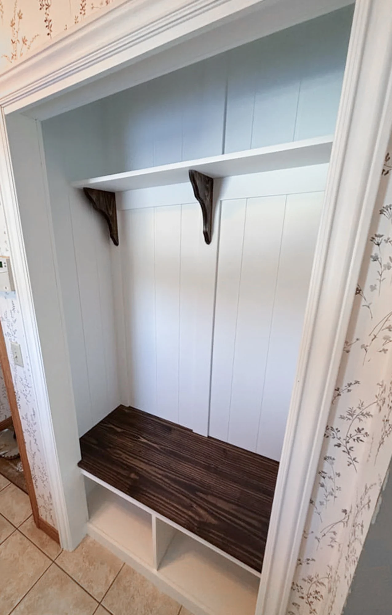 Empty built-in closet with a single shelf and a dark wood bench, with two small storage cubbies below, and floral wallpapered wall on the right.