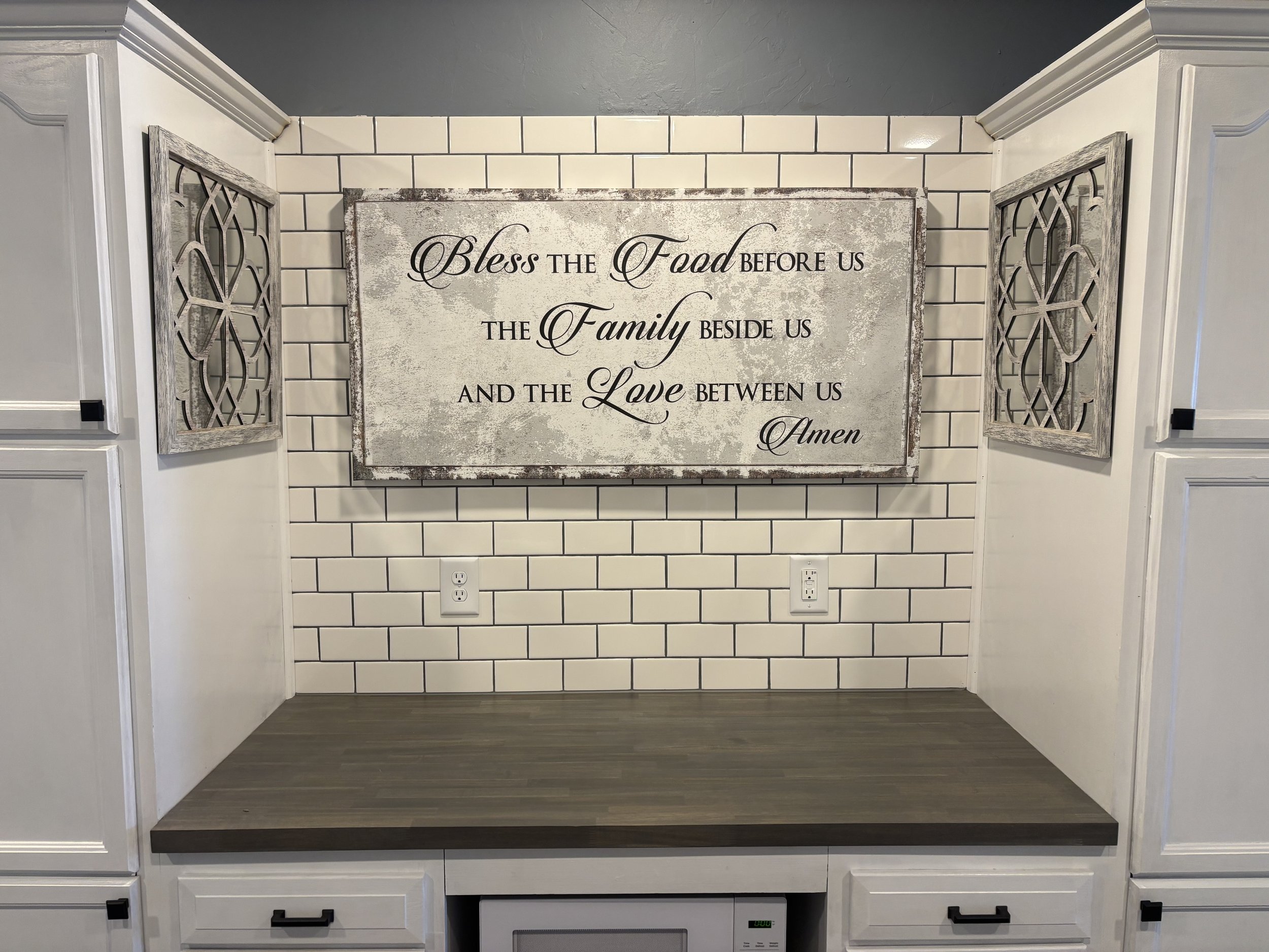 Kitchen wall with a sign that reads, 'Bless the Food before us, the Family beside us, and the Love between us. Amen,' flanked by two decorative wooden panels and surrounded by white cabinets.
