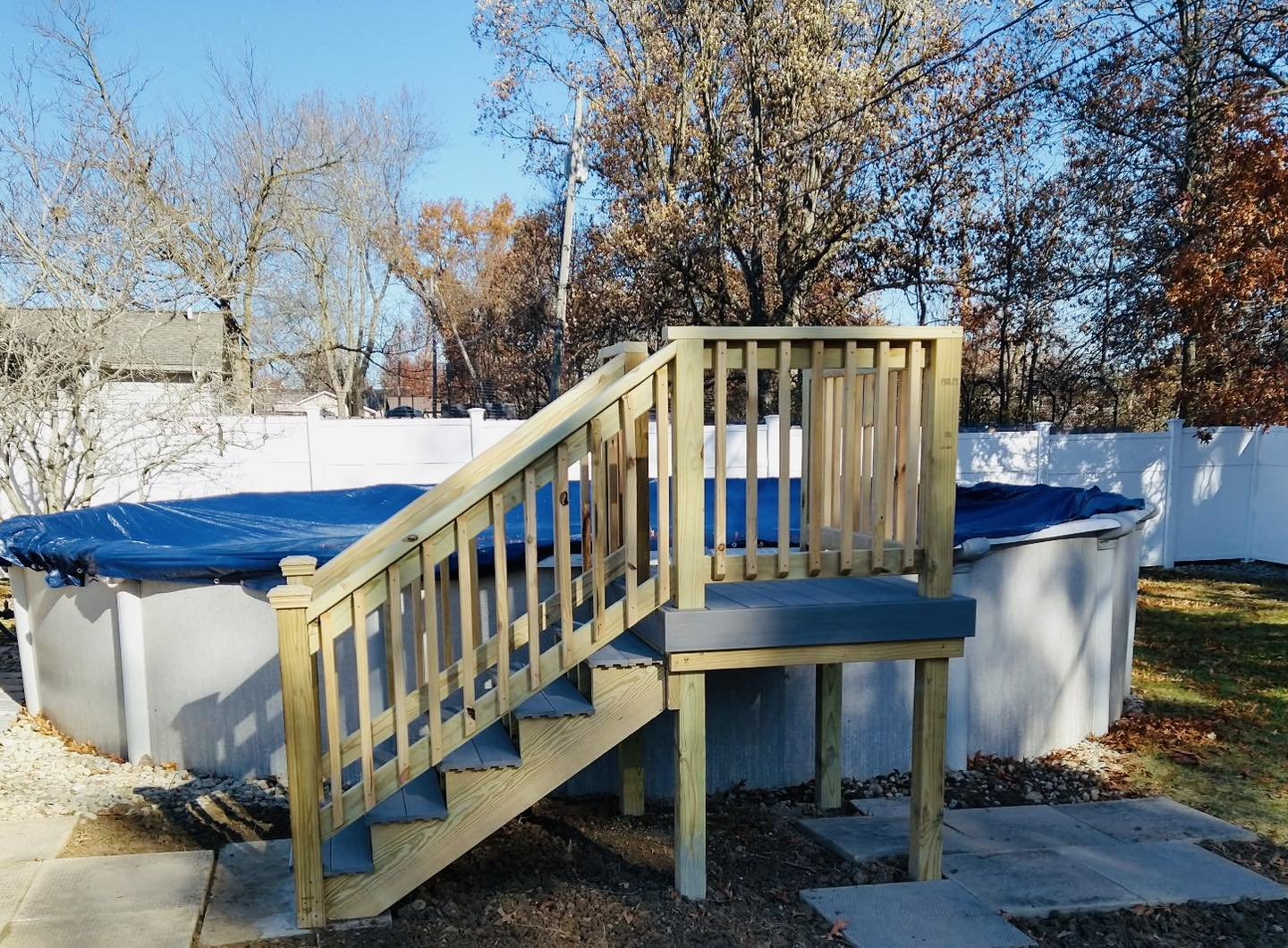 An above-ground pool with a blue cover and a new wooden staircase and railing leading up to it, situated in a backyard with a white fence and trees with autumn leaves in the background.