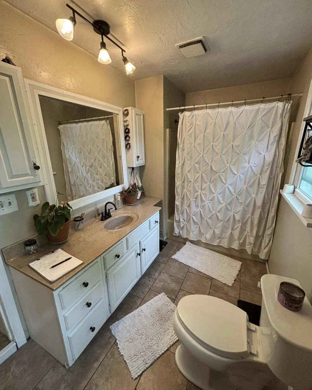 Bathroom with beige walls and tiled floor, white vanity with a sink, large mirror, potted plants, and white curtains on a shower or bathtub area. There are two white bath mats, a toilet, and a window.