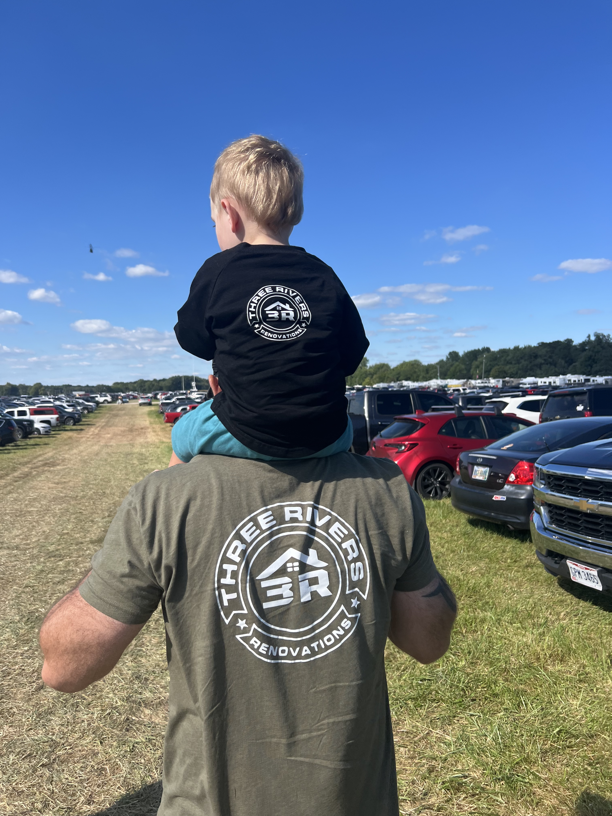 A man is holding a child on his shoulders while walking through a parking lot on a clear, sunny day. Both are wearing t-shirts with the 'Three Rivers Renovations' logo.