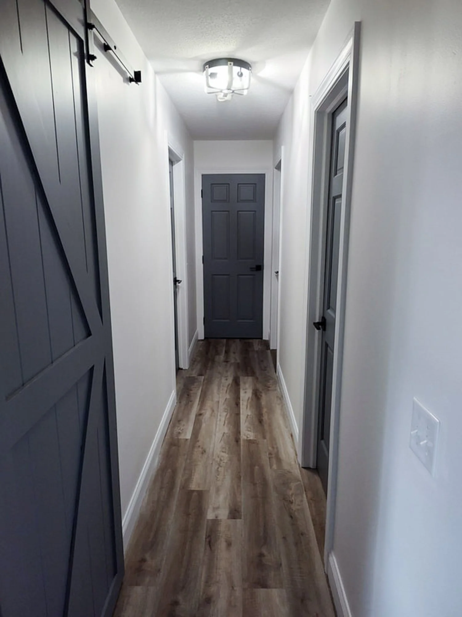 A narrow hallway with white walls, multiple gray doors with black handles, a light fixture on the ceiling, and wooden flooring.