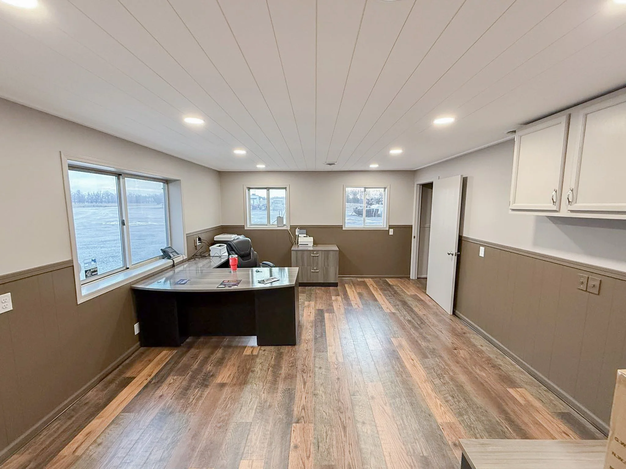 An empty office room with wood flooring, white and brown walls, overhead lights, two windows, and a desk with office supplies.