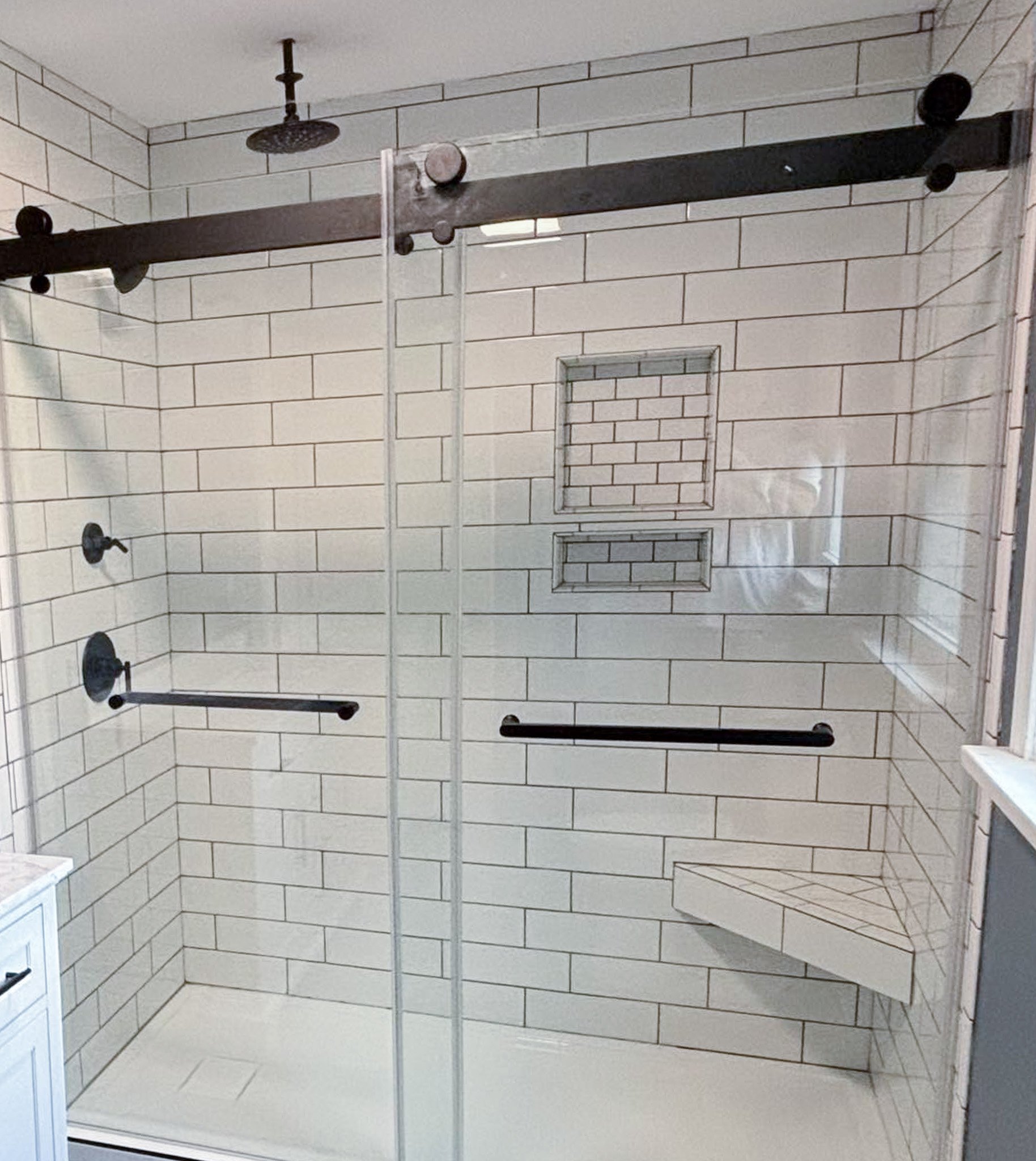 Modern shower with white subway tile walls, a black sliding barn-style door, two black shower knobs, a black shower head, a built-in tiled niche, and a corner tiled bench.