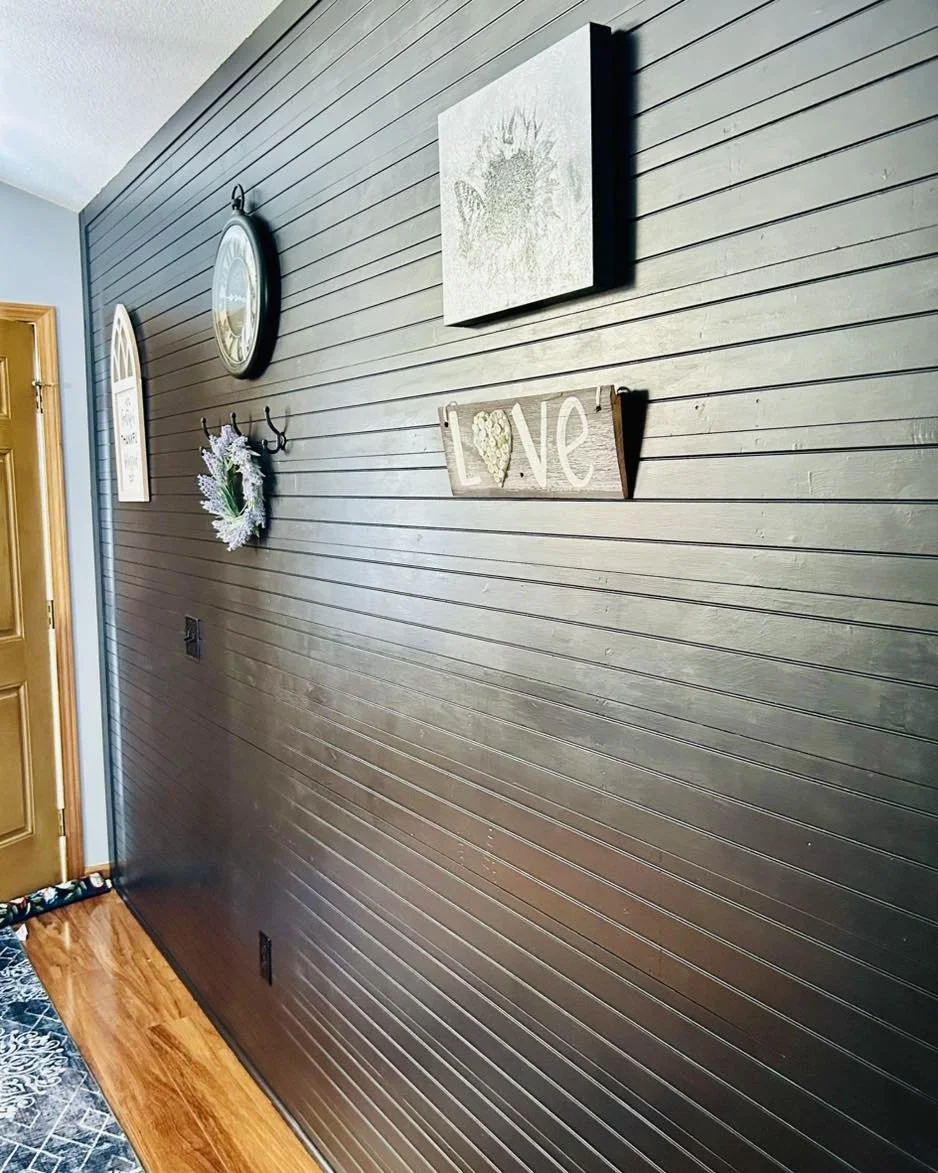 Decorated black wall with wall art, clock, and hooks. Wooden door on the left, wooden floor, and a black and white rug at the bottom.