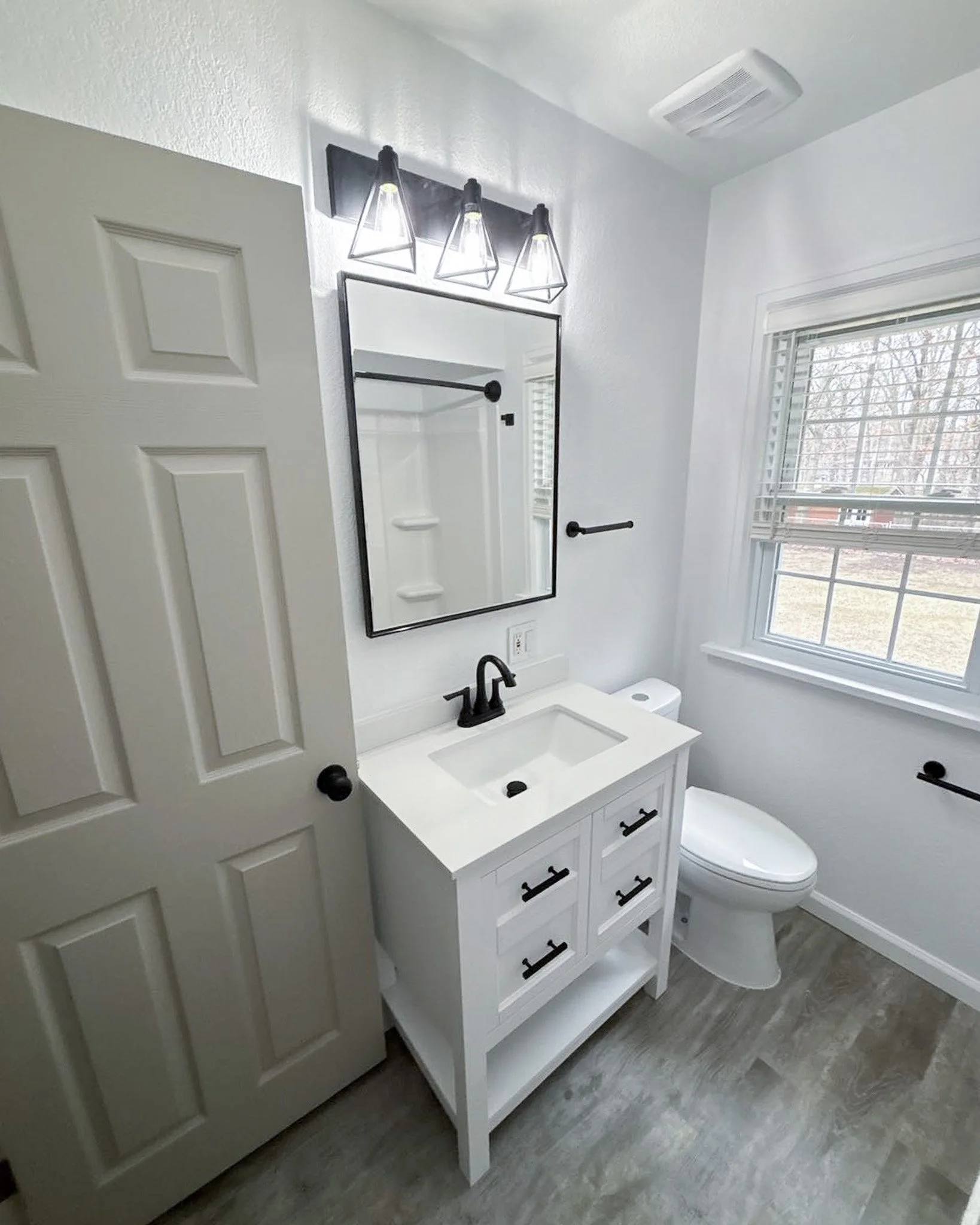 A modern white bathroom with a vanity, black fixtures, a mirror, and a window with blinds.