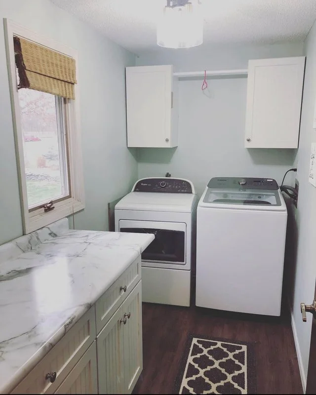 Laundry room with a window, white cabinets, a marble countertop, a front-loading washer, and a top-loading dryer, with a patterned rug on dark wood floor.
