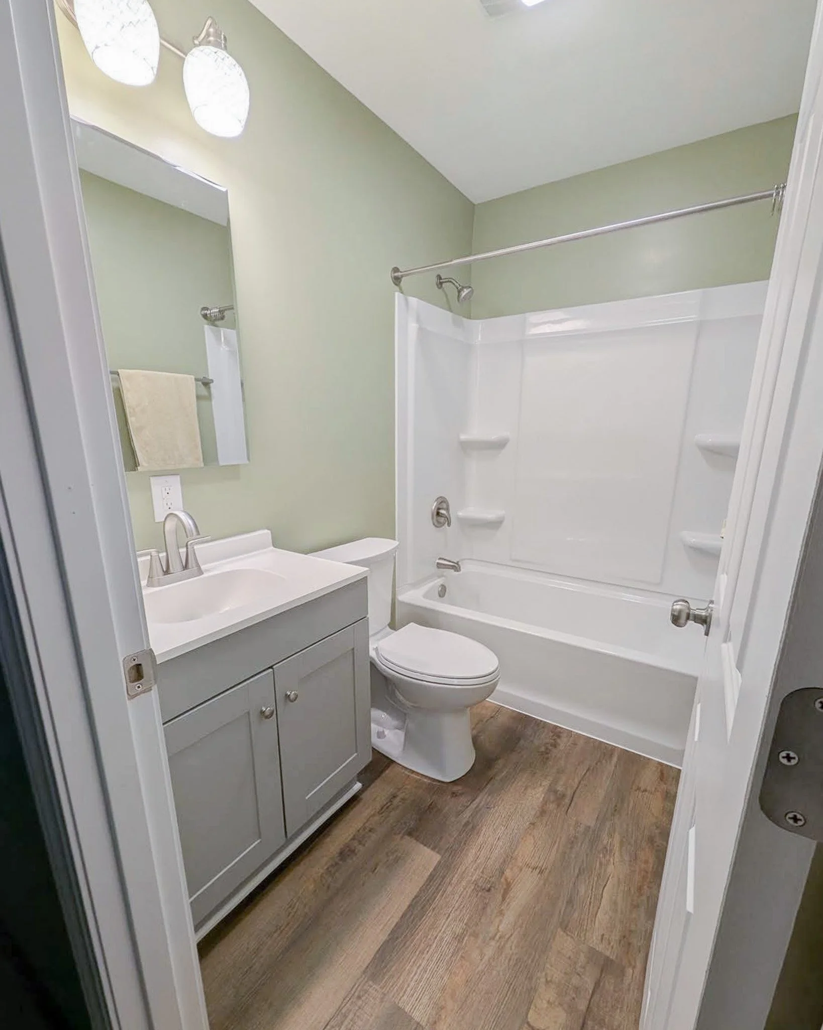 A small bathroom with a white vanity, mirror, and toilet next to a bathtub with a shower curtain rod. The walls are light green, and the floor has wood-like tiles.