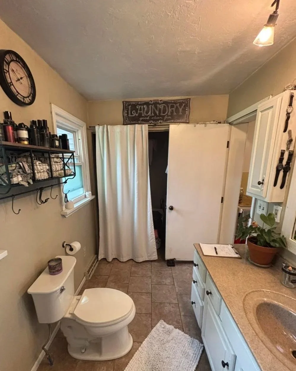 A small bathroom with beige walls, a toilet, a window, a shelf with toiletries, a wall clock, a bathroom rug, a vanity with a plant and a clipboard, a tile floor, and a closet with a white curtain.
