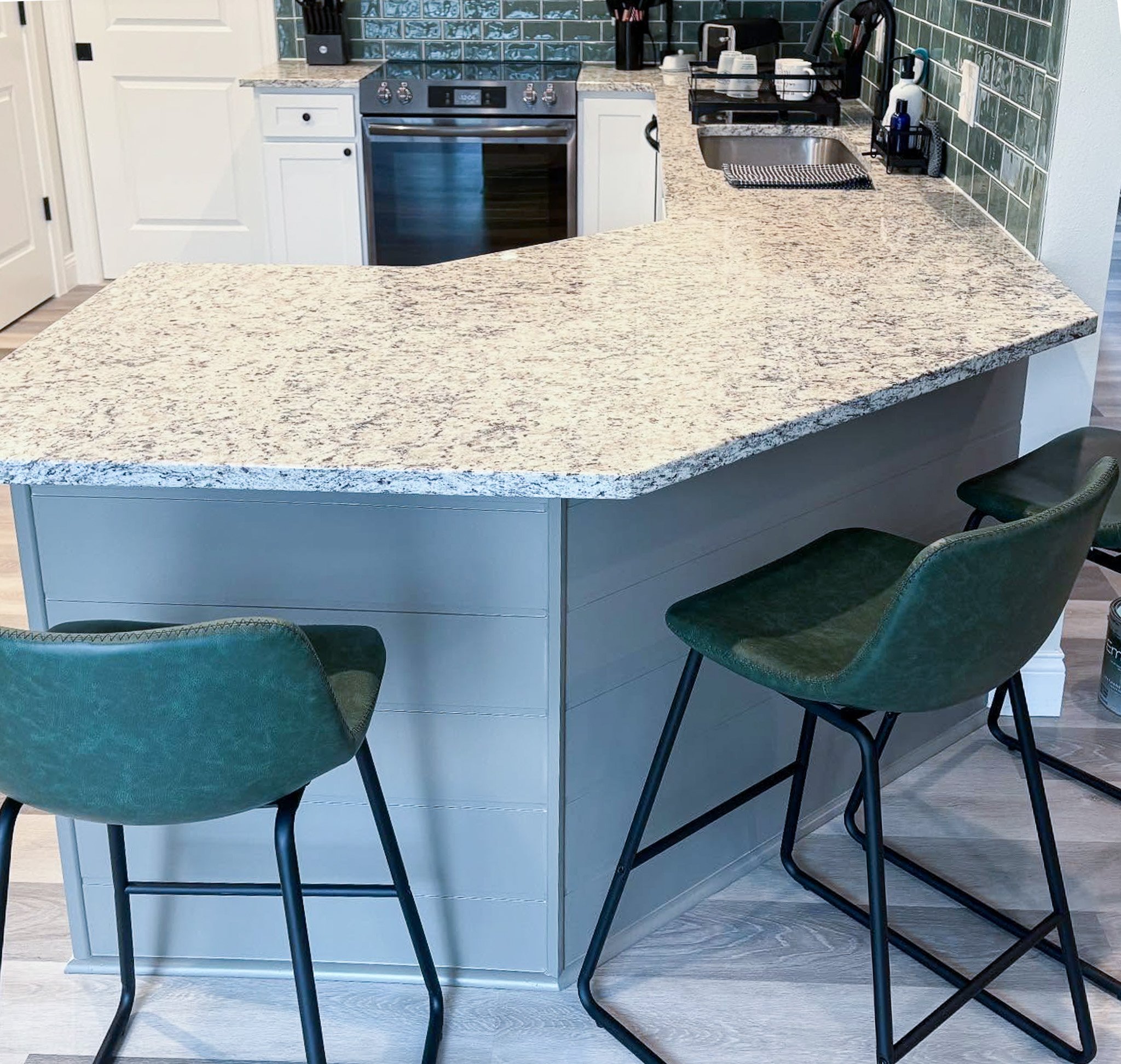 Kitchen island with granite countertop, two green bar stools, and white cabinets. In the background, there is a stove, backsplash tiles, and a small sink area with dish drying rack.