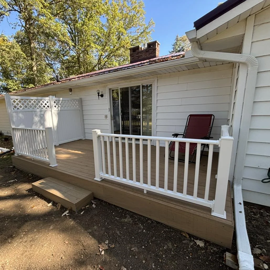 New wooden porch on a white house with a sliding glass door, white railings, a red outdoor chair, a small set of stairs, and a background of trees and a blue sky.