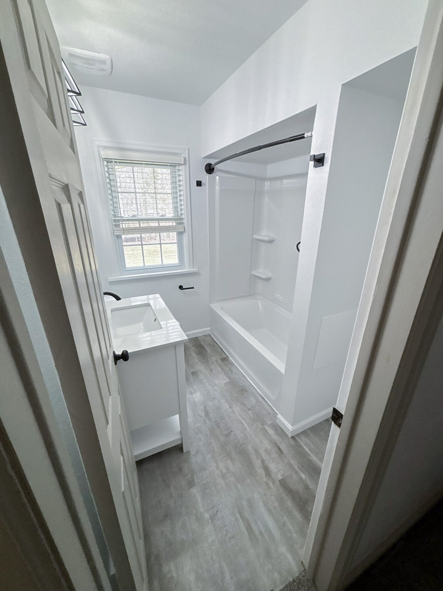 A small bathroom with a window, vanity, bathtub with shower, and grey wood-patterned flooring.