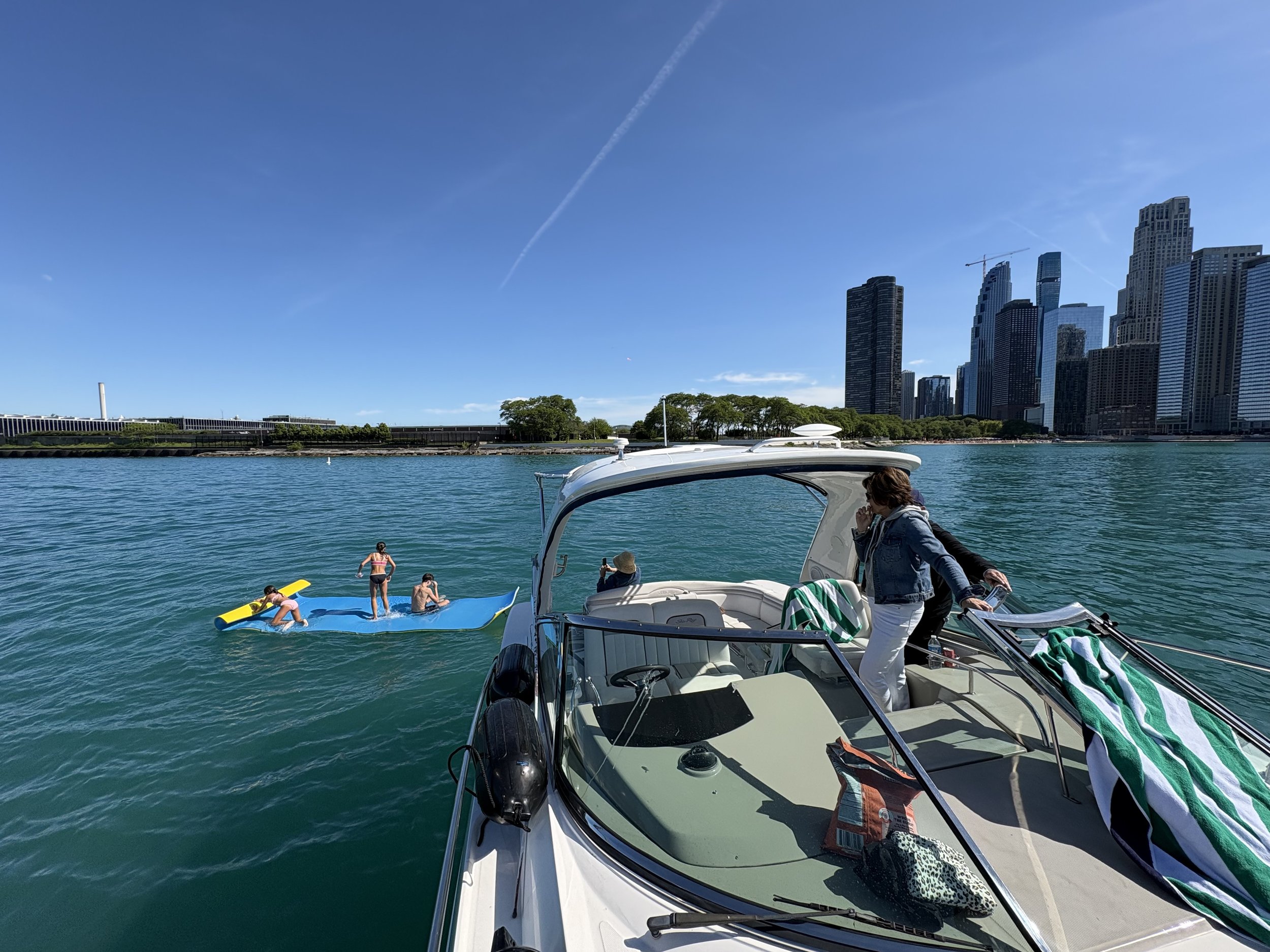 People enjoying water activities near a city skyline, with a boat in the foreground and tall buildings in the background, and a clear blue sky.