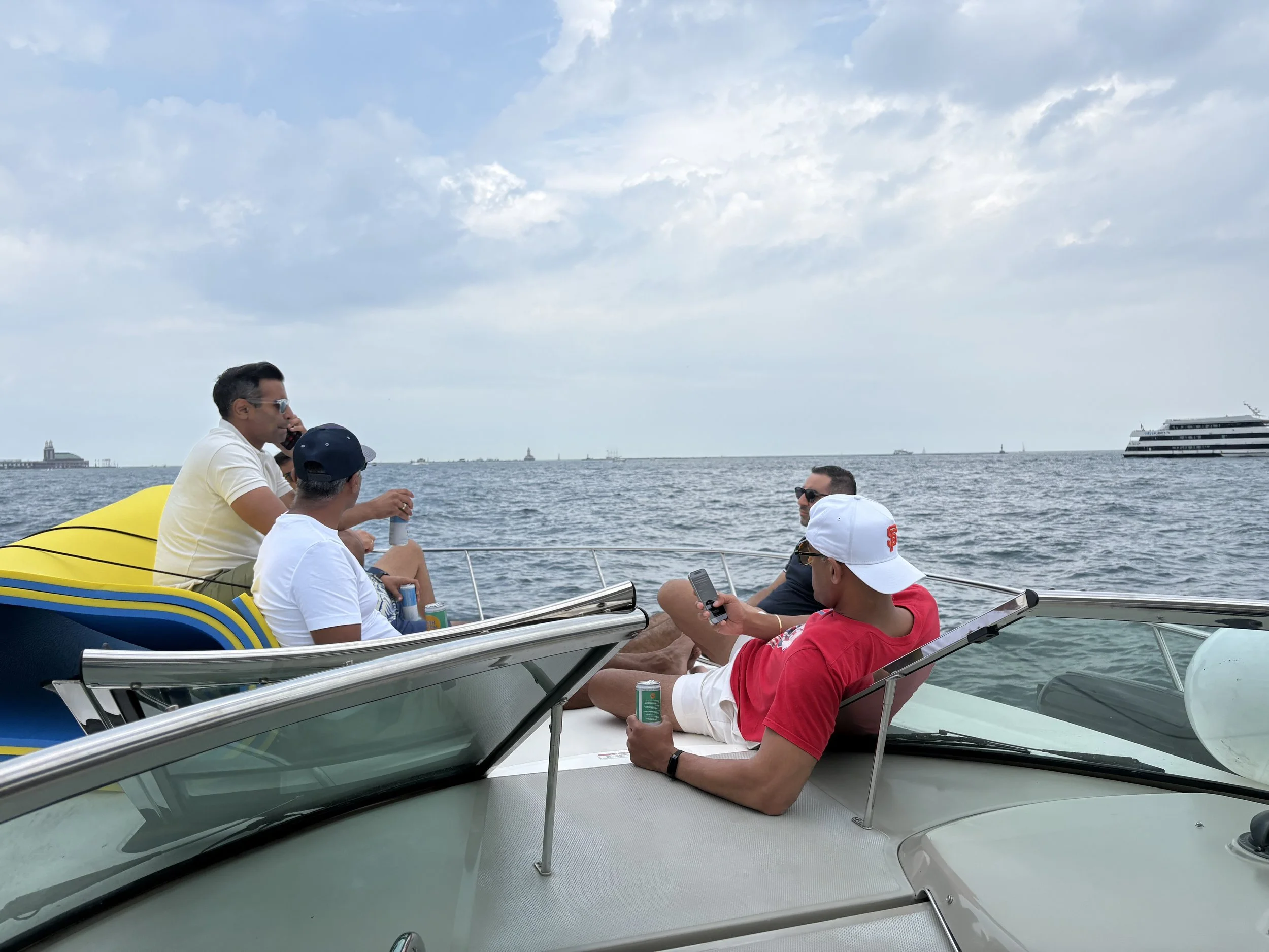 Group of people sitting on a boat, relaxing and talking, with a large cruise ship and the ocean in the background.