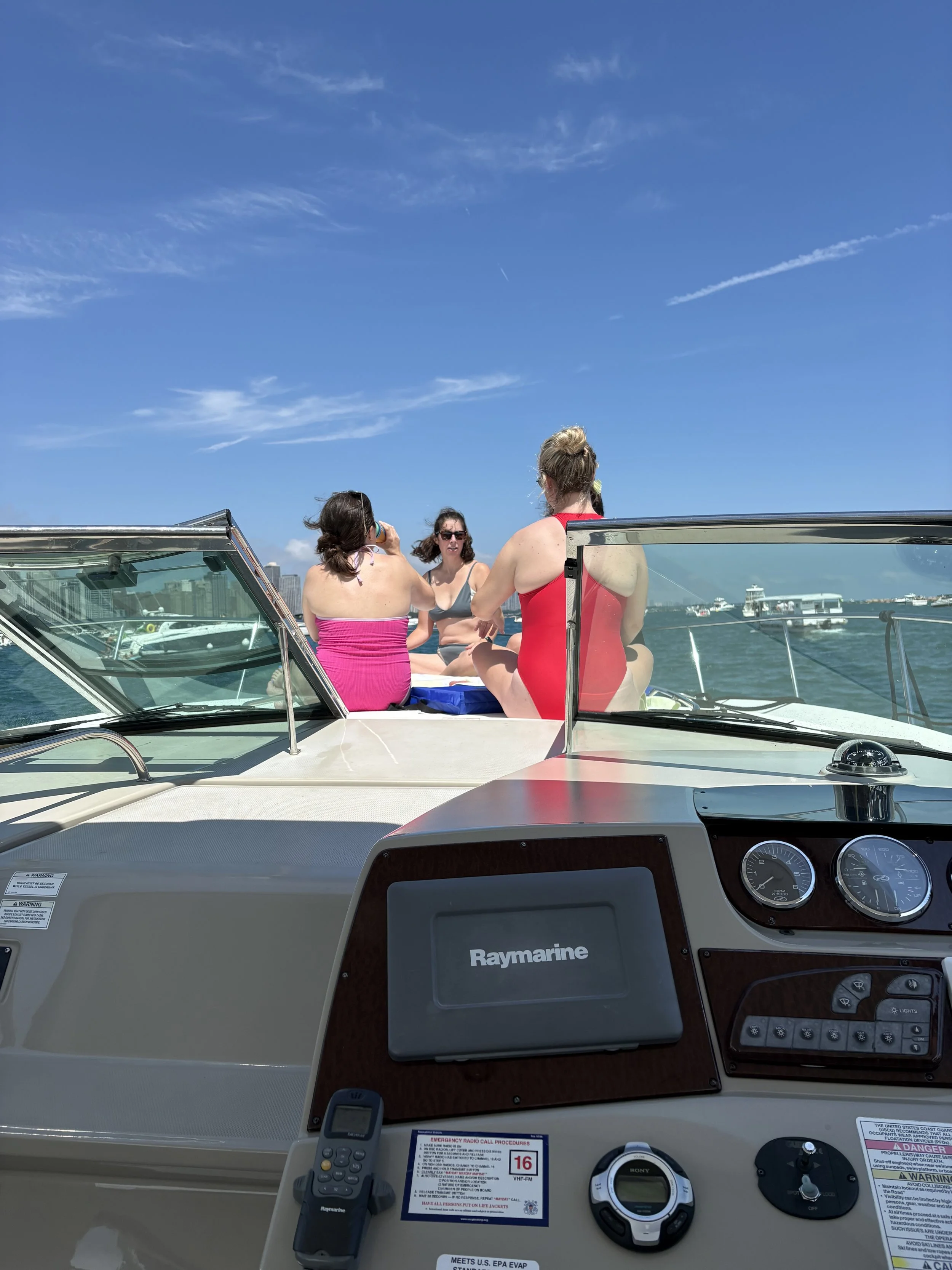 View from the boat showing three women on a boat deck, one drinking from a cup, two facing away, with a city skyline in the distance and a blue sky with clouds.
