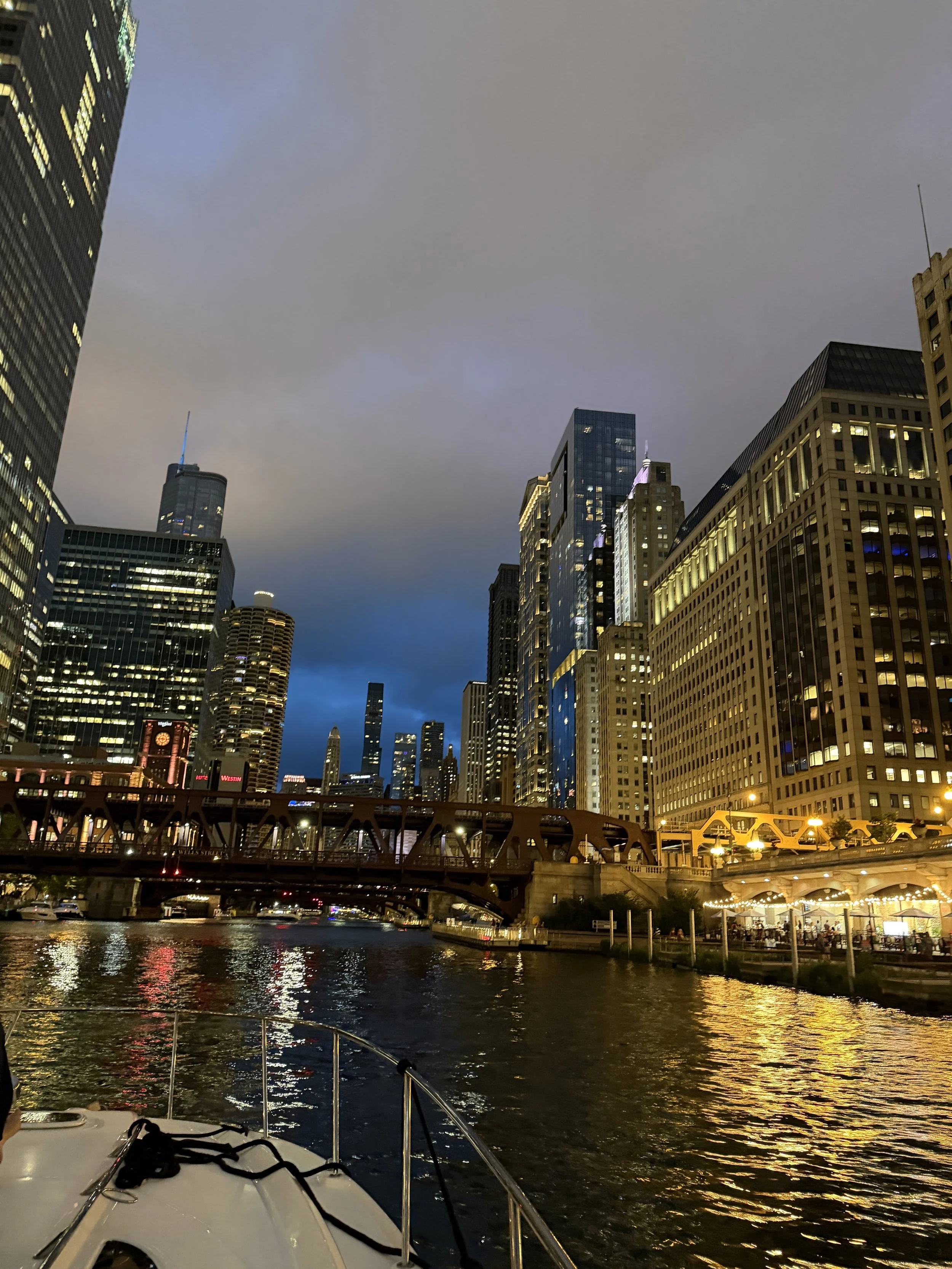 Nighttime cityscape with tall buildings, bridge over water, and boat in the foreground, illuminated windows and reflections in the water.