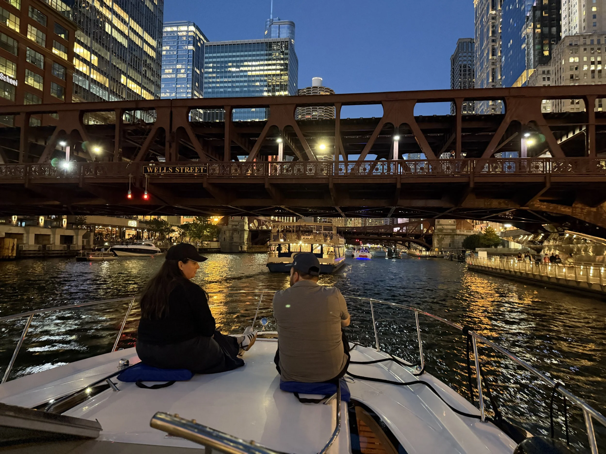 Two people sitting on a boat on a city river at dusk, with tall modern buildings lit in the background and a bridge overhead.