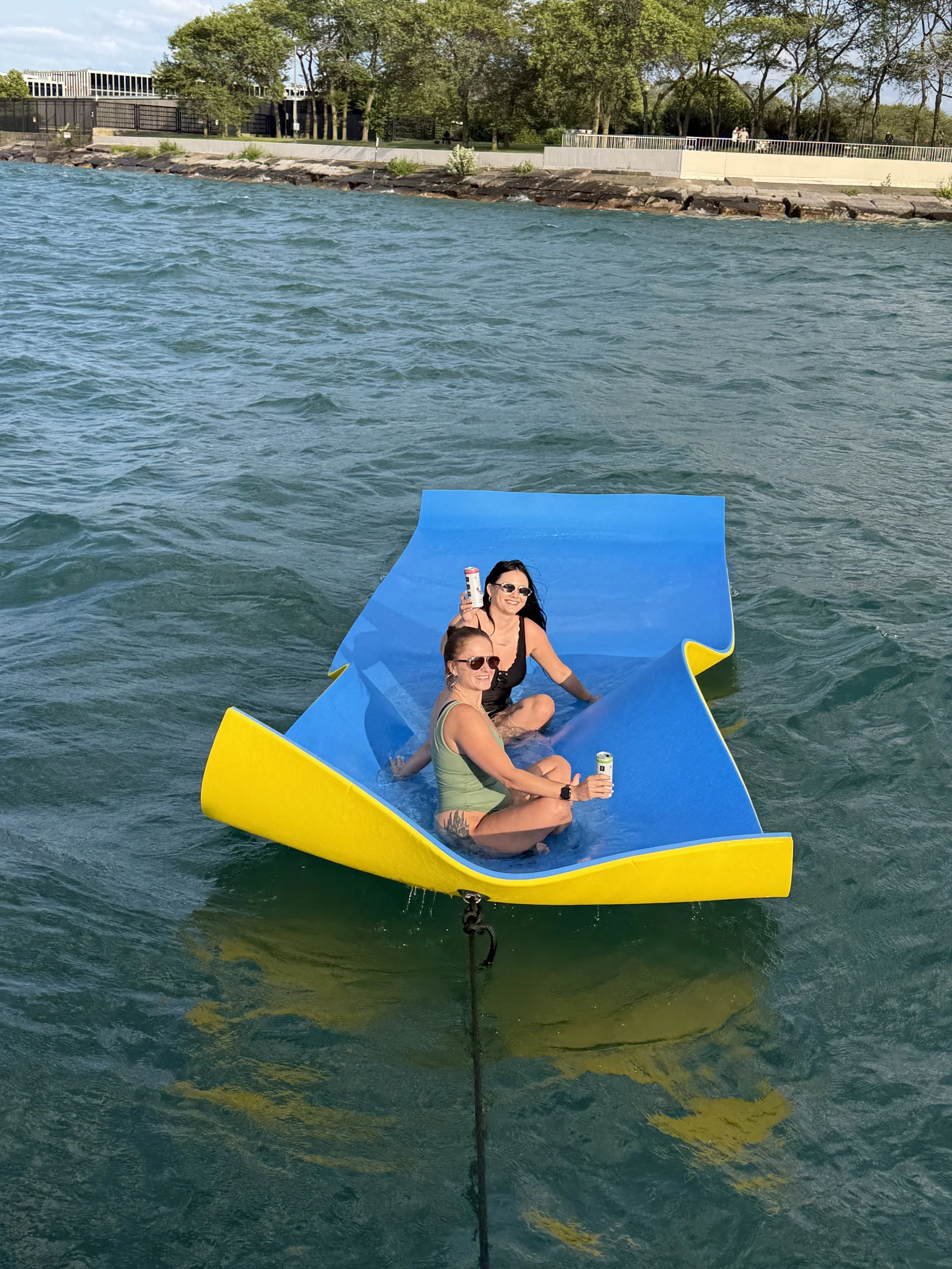 Two women sitting on a floating device on the water, one holding a drink and both smiling, with trees and a walkway in the background.