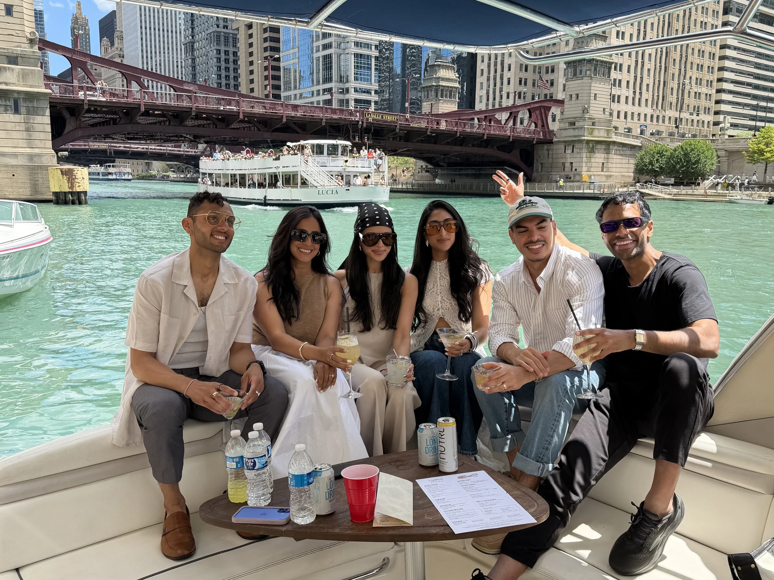 A group of six friends sitting on a boat with a city skyline and a bridge in the background, enjoying drinks and smiling at the camera during daytime.