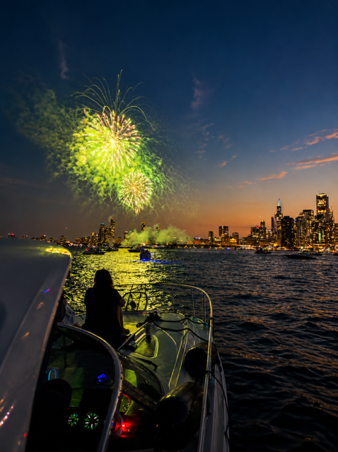 A person sitting on a boat watching fireworks over the city skyline at dusk.