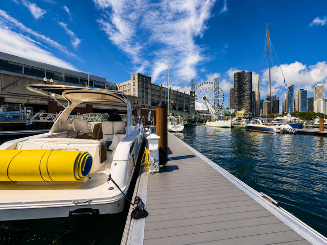 A marina with yachts docked, a city skyline with high-rise buildings, a Ferris wheel, and a blue sky with clouds.