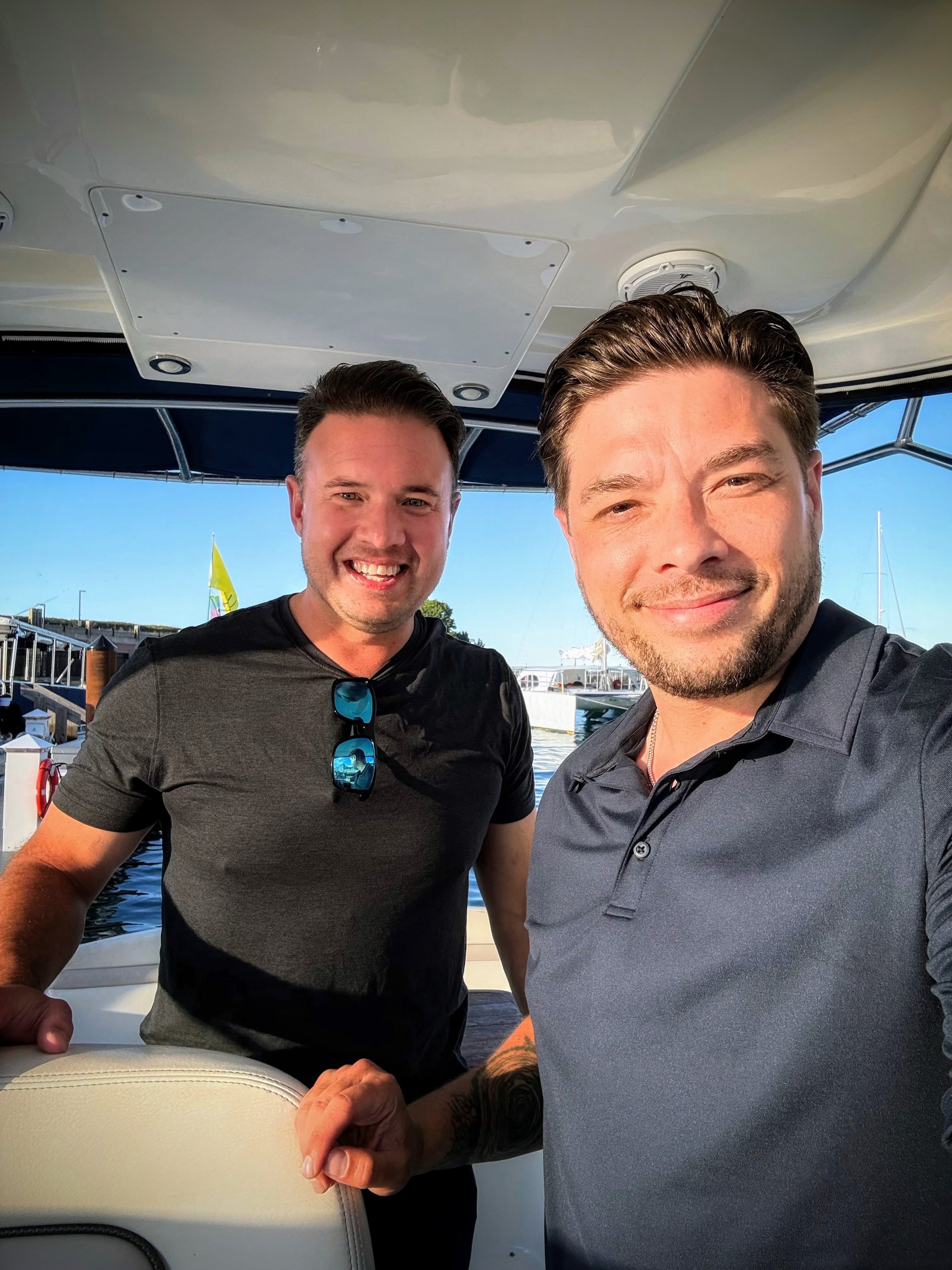 Two smiling men taking a selfie on a boat dock, with boats and flags visible in the background, and one man showing his tattooed arm.