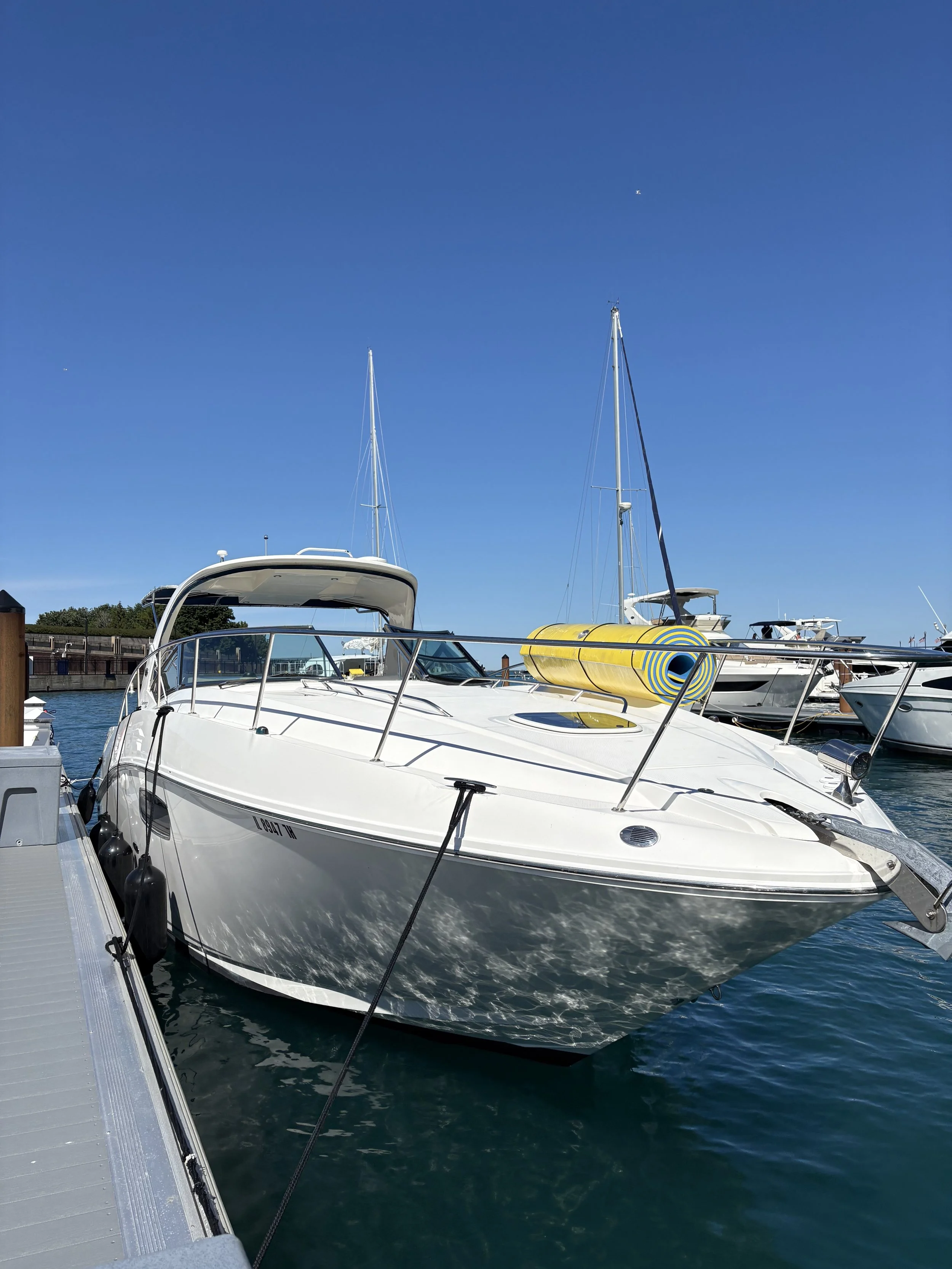 A white motorboat docked at a marina with sailboats in the background under a clear blue sky.