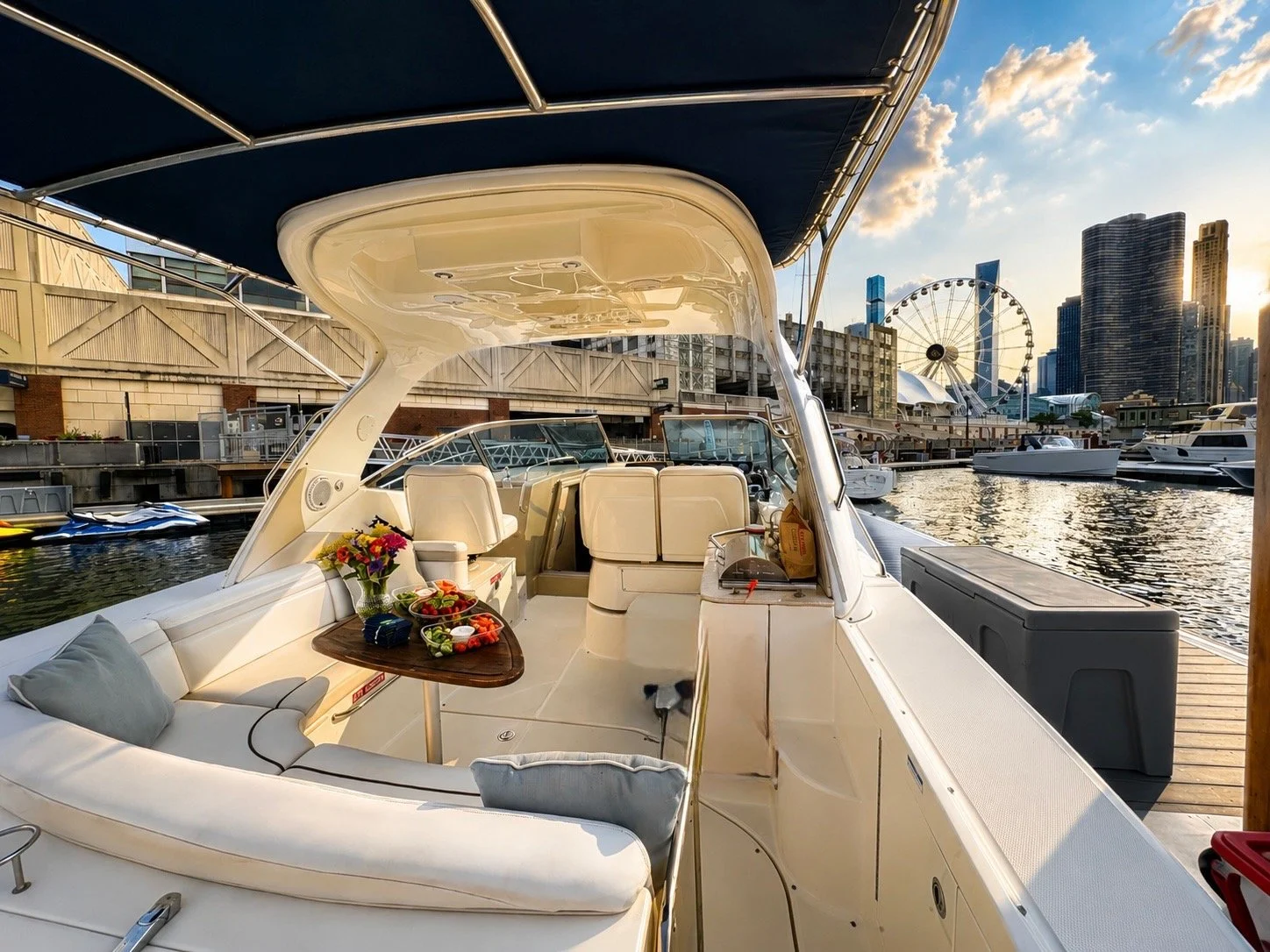 A yacht docked at a marina during sunset with a city skyline, including a Ferris wheel, in the background. The yacht's deck has a table with fruit and a flower vase, and cushioned seating.