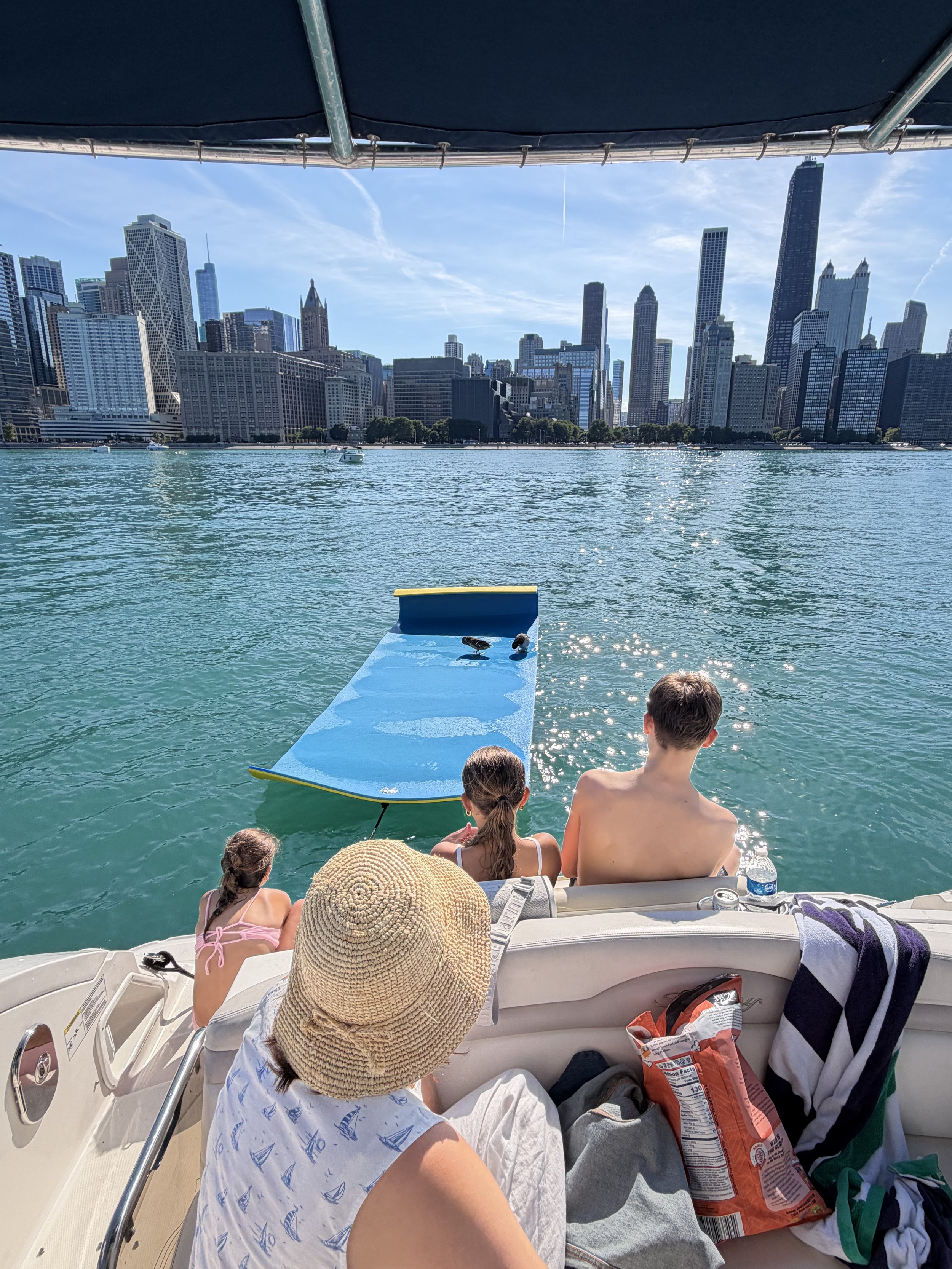 People relaxing on a boat, sitting on the deck, with a city skyline in the background and a clear blue sky.