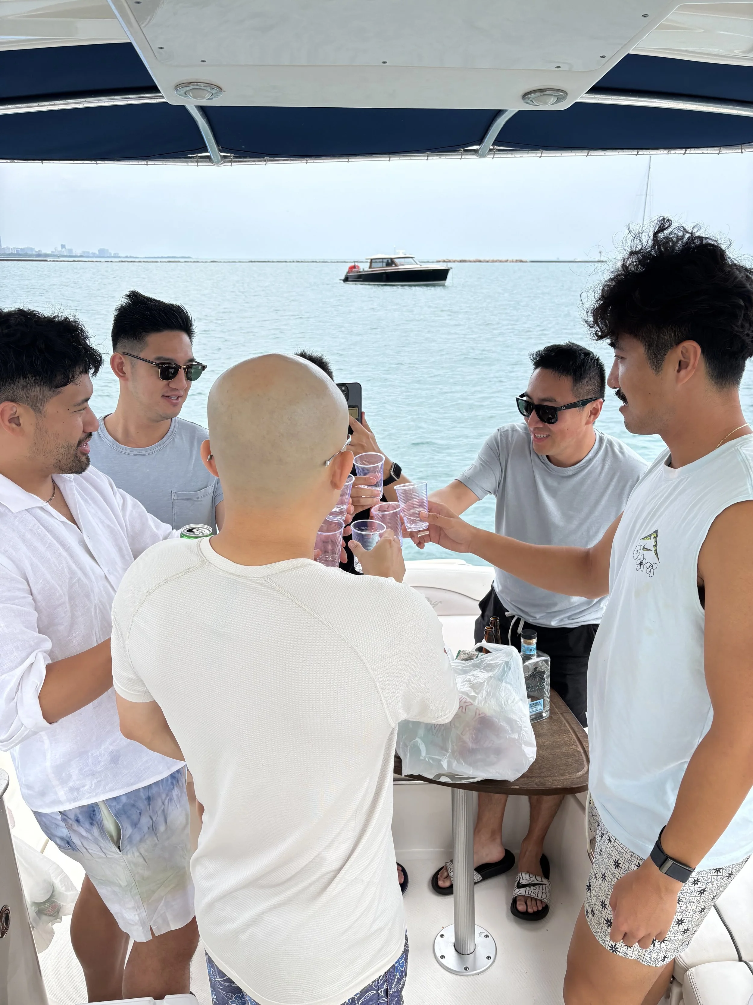 A group of six people with diverse skin tones on a boat sharing a toast with plastic cups while enjoying a day out on the water. The background shows calm water, a boat, and a distant skyline.