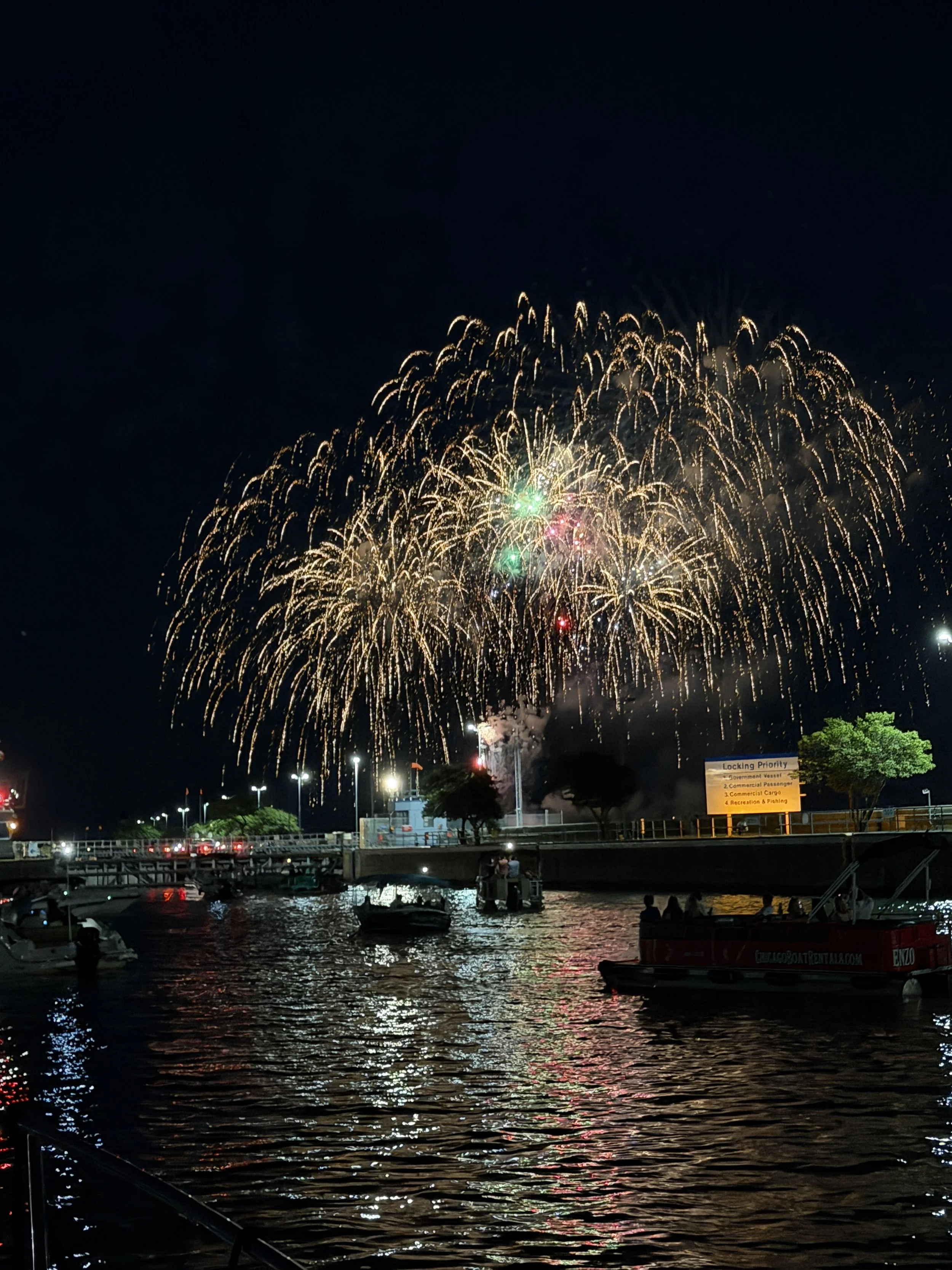 Fireworks display over a body of water with boats and a bridge, illuminated by various lights at night.