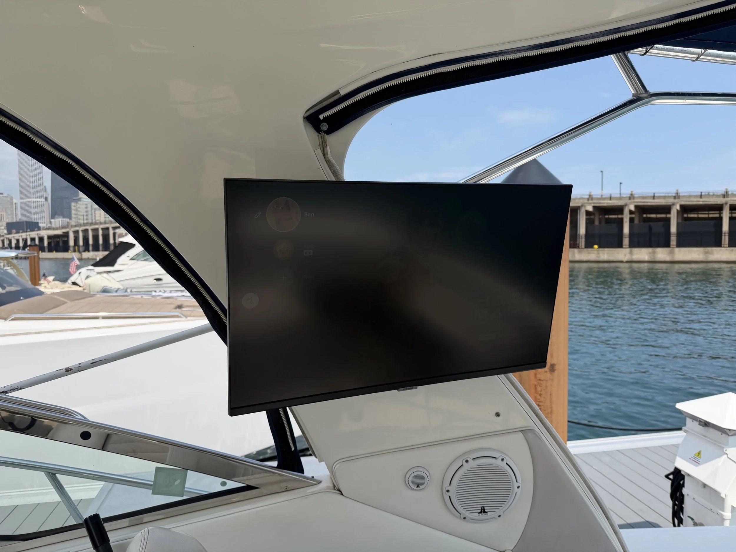 Close-up view of a yacht's interior showing a flat-screen monitor, speakers, and part of the boat's ceiling with window and city skyline in the background.