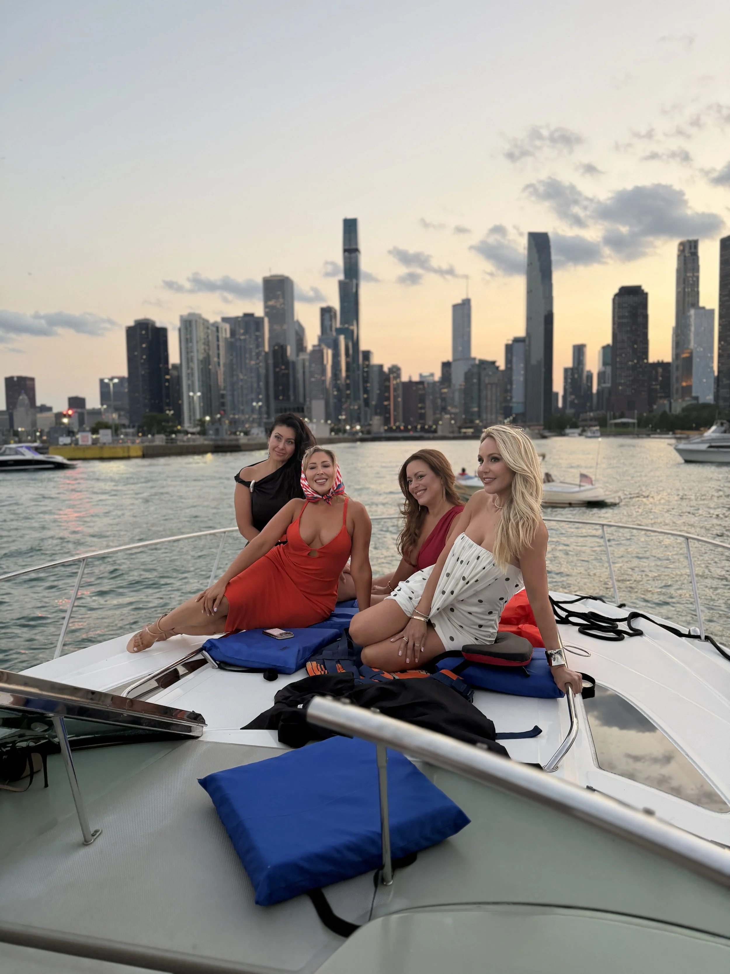 Four women sitting on a boat during sunset with a city skyline and boats in the water behind them.