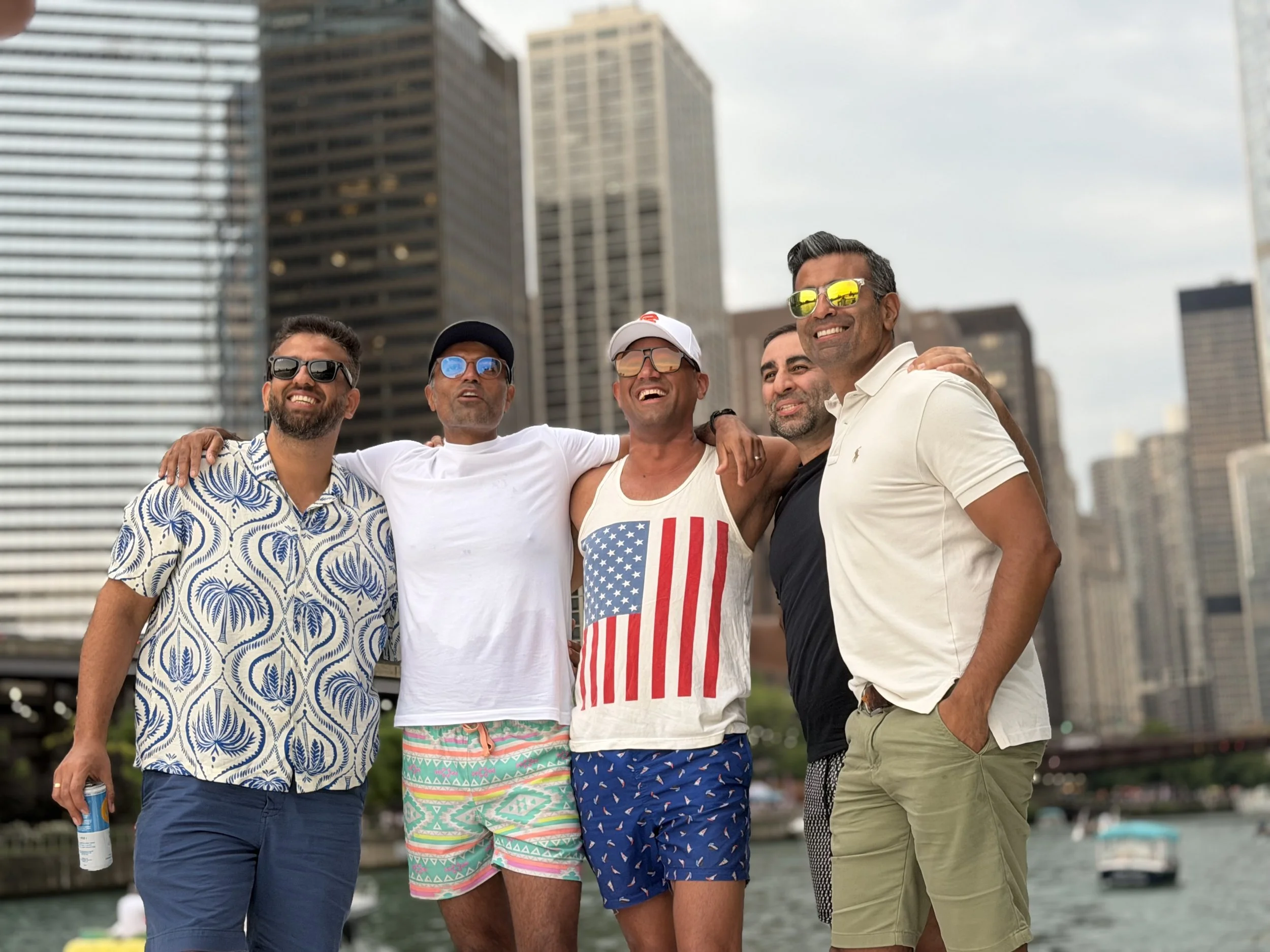 A group of five men standing closely together on a city rooftop with tall buildings in the background, smiling and enjoying each other's company in casual summer clothes and sunglasses.