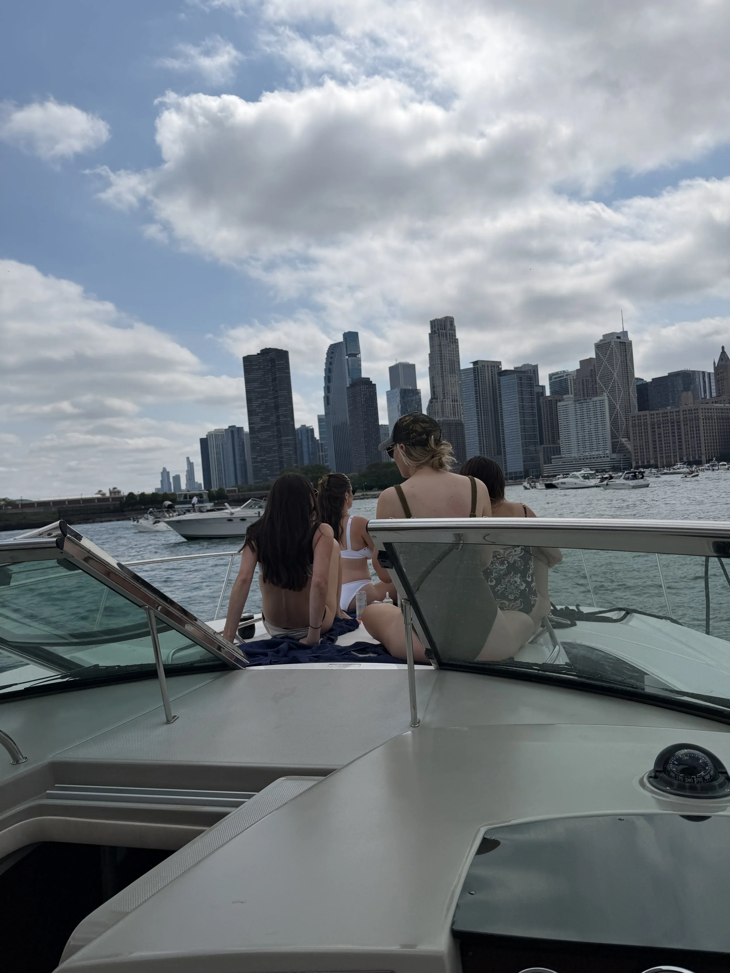 Four women sitting on a boat with sunglasses, talking, with a skyline of tall buildings and a partly cloudy sky in the background.