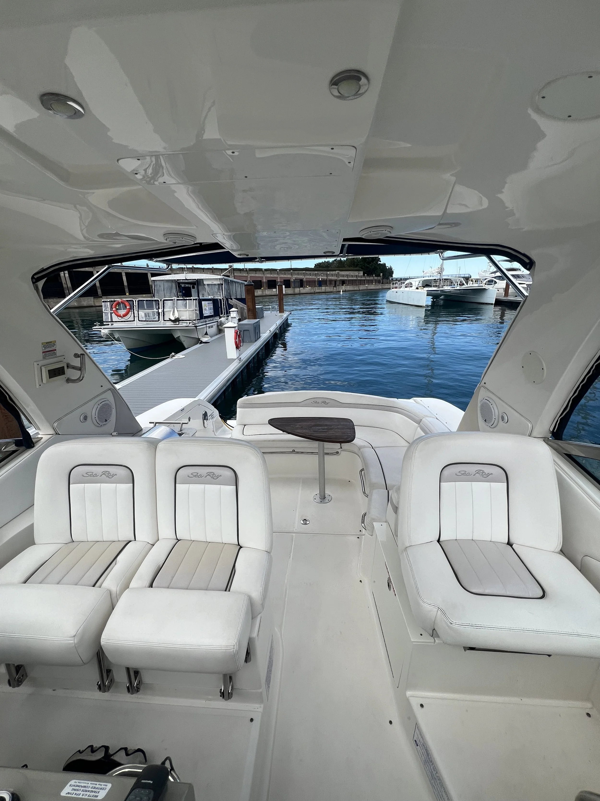 Interior view of a boat showing white cushioned seating with headrests, a small table, and the view through an opening to the dock with boats and water outside.