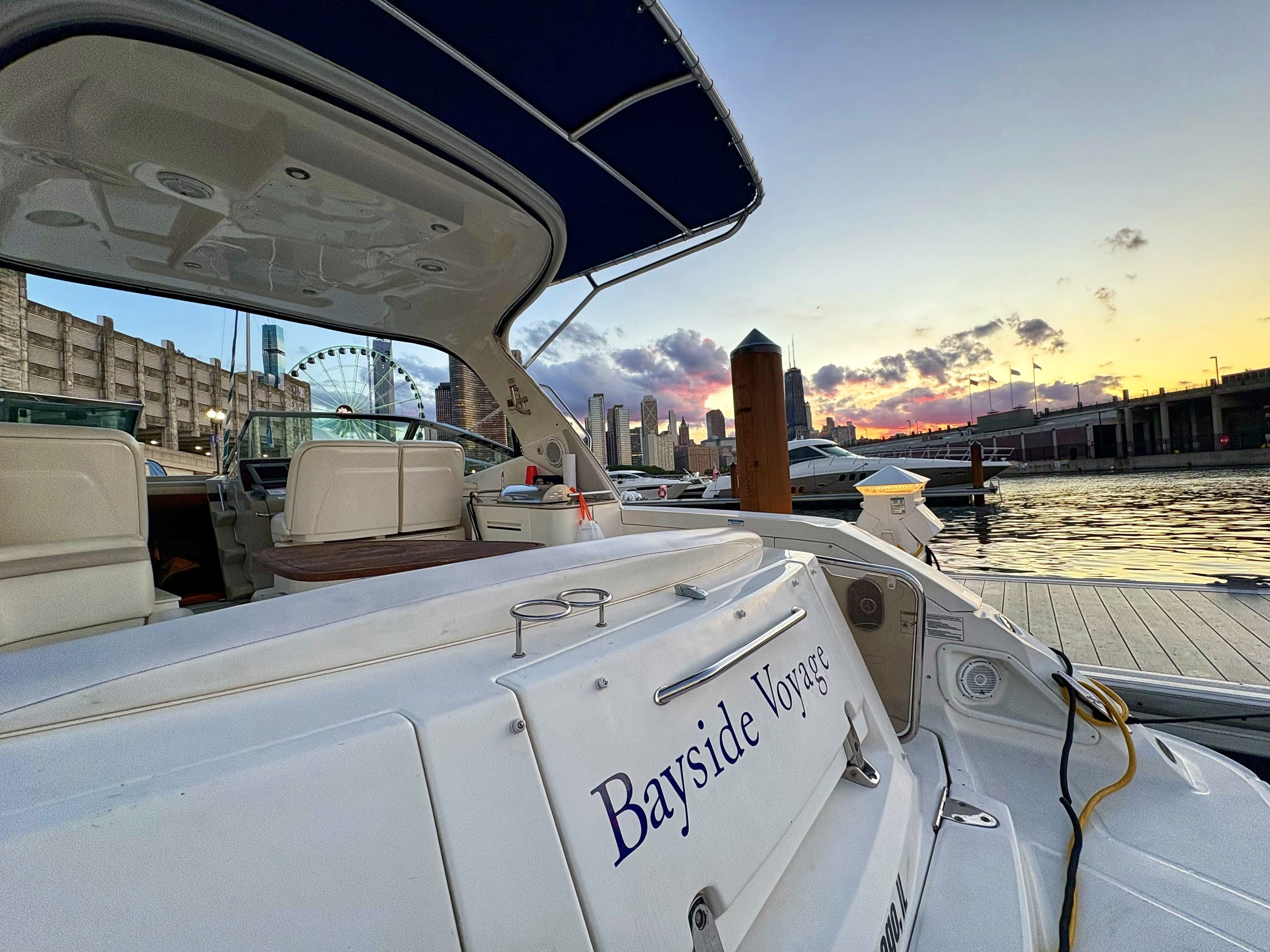 View from a docked boat showing part of its roof, a city skyline with tall buildings, a ferris wheel, and a sunset sky with clouds and warm colors.