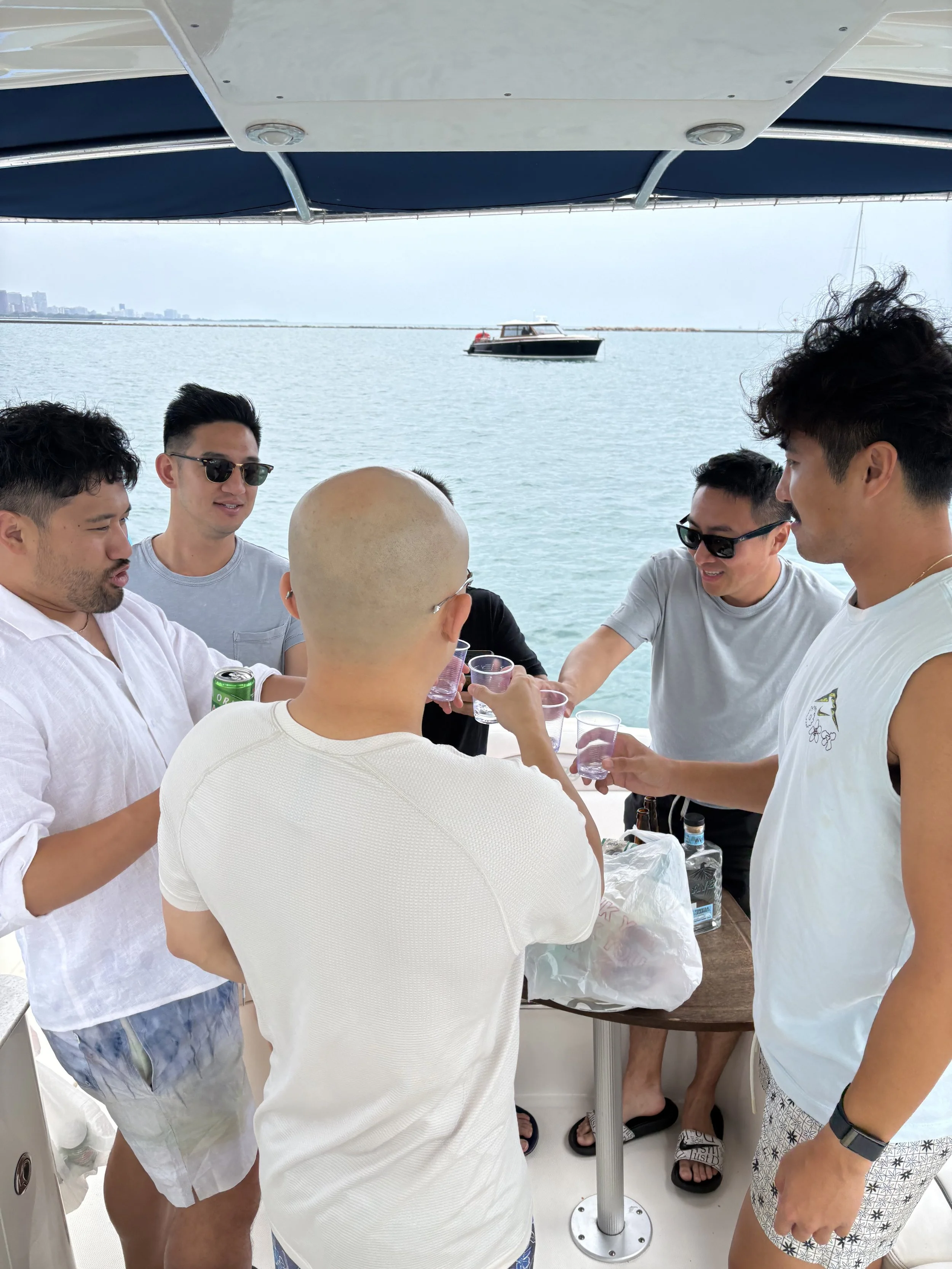Group of people on a boat holding small cups, with a body of water, a boat, and a distant shoreline in the background.