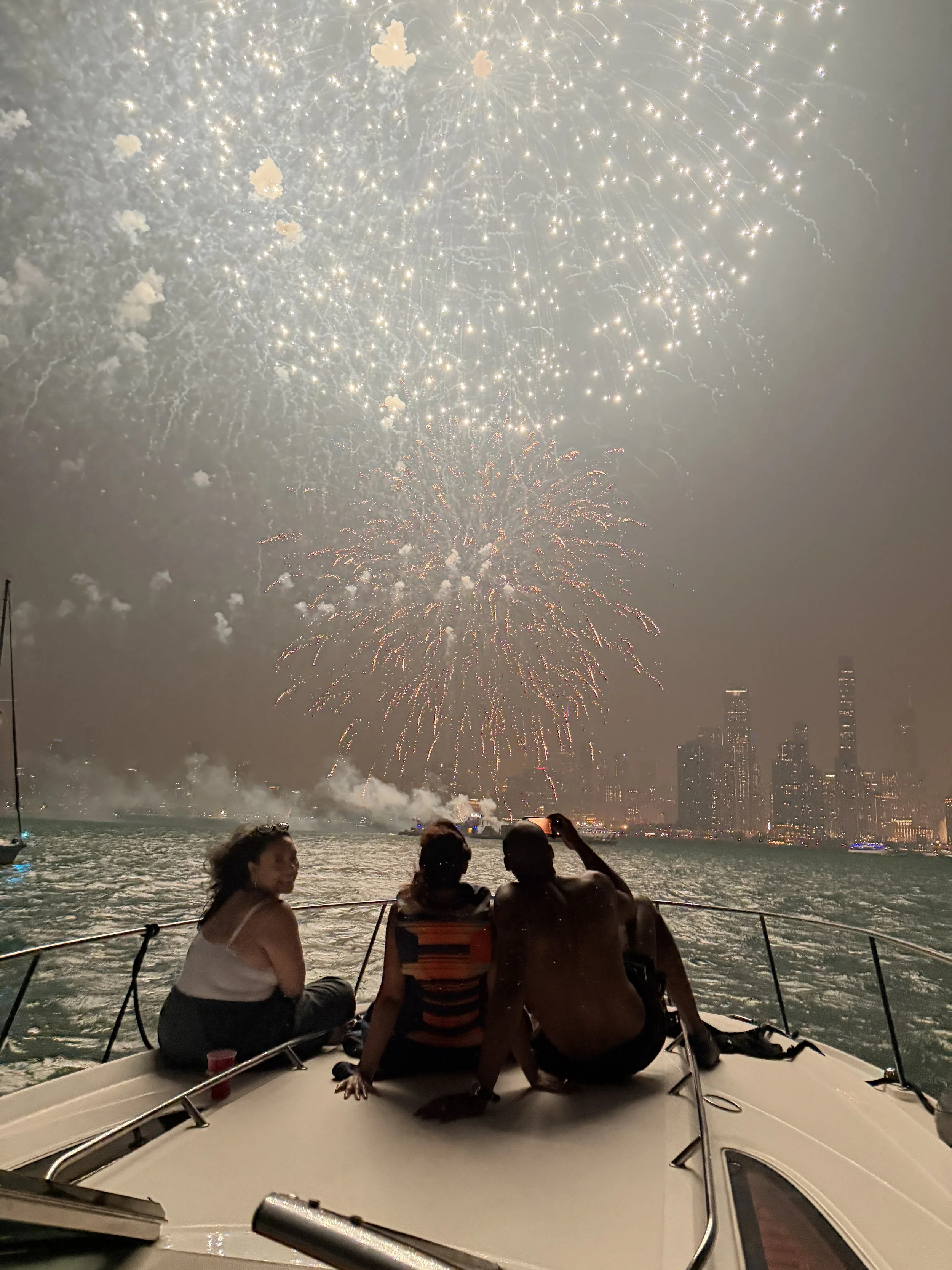 People sitting on a boat watching fireworks light up the night sky over a city skyline with tall buildings.