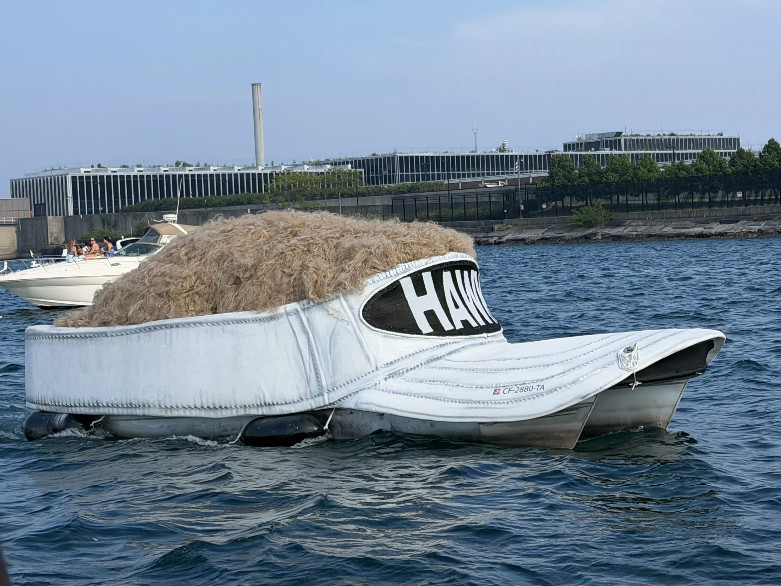A close-up view of a white inflatable boat with a tan furry object on top, floating on blue water near a shoreline with trees, modern buildings, and boats in the background.