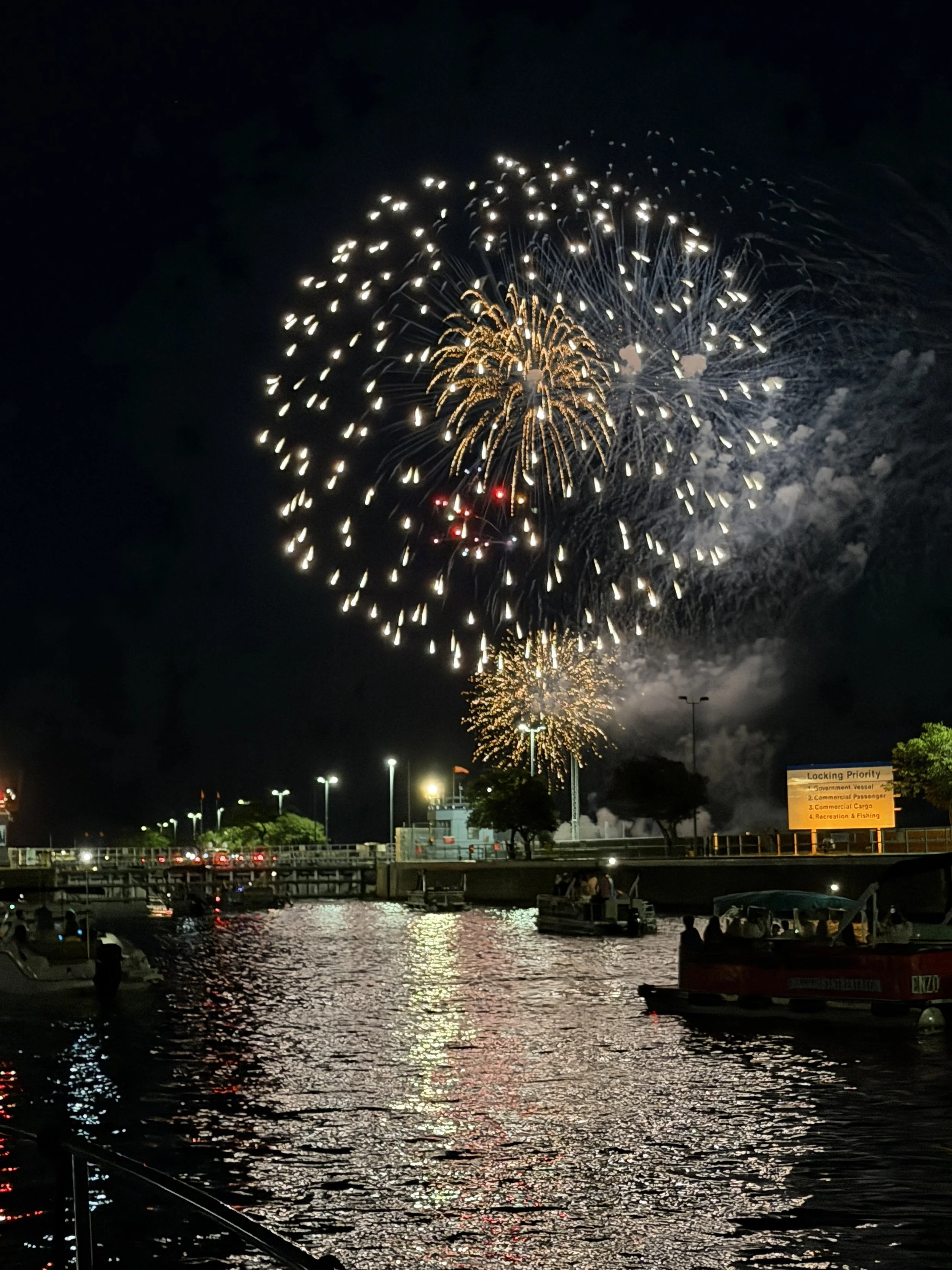Nighttime fireworks display over a harbor with water, boats, and a dock. Bright fireworks light up the dark sky with reflections on the water, and a sign visible in the foreground lists locking priority information.