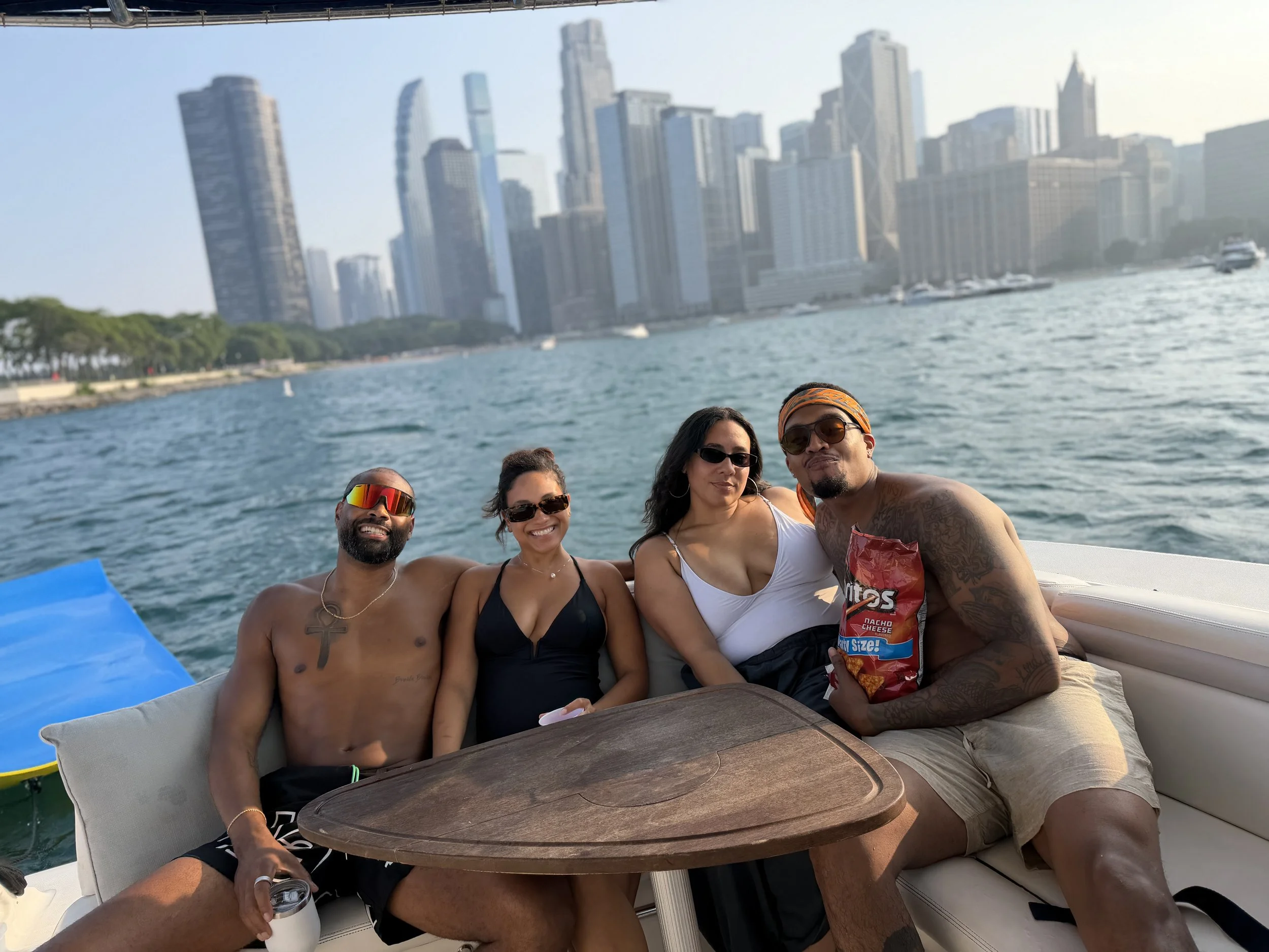 Five people sitting together on a boat in a city waterfront, with a skyline of tall buildings in the background, smiling and enjoying the sunny day.