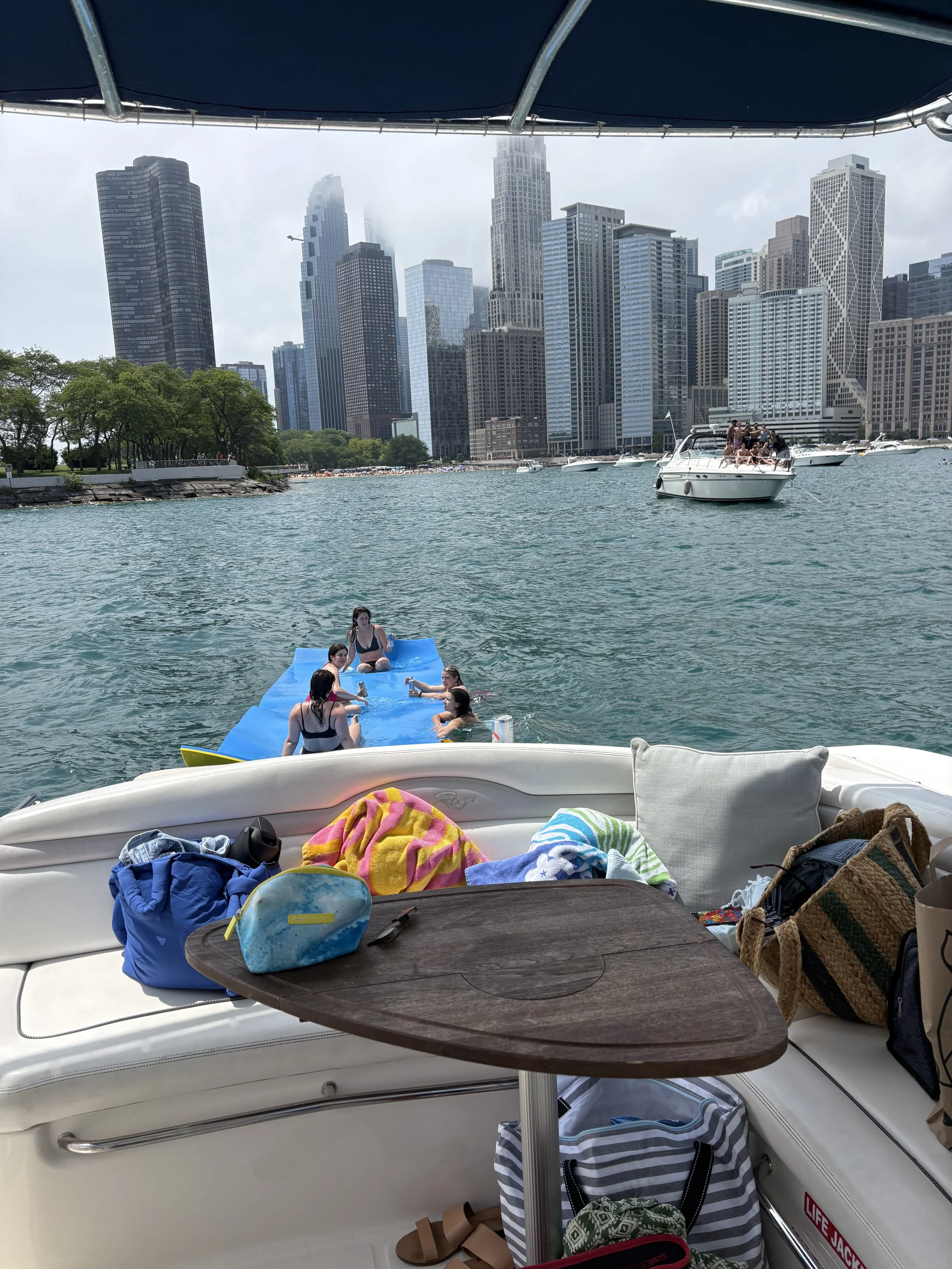 View from a boat showing a city skyline with tall skyscrapers, boats in the water, and people relaxing on a boat with towels, bags, and drinks.