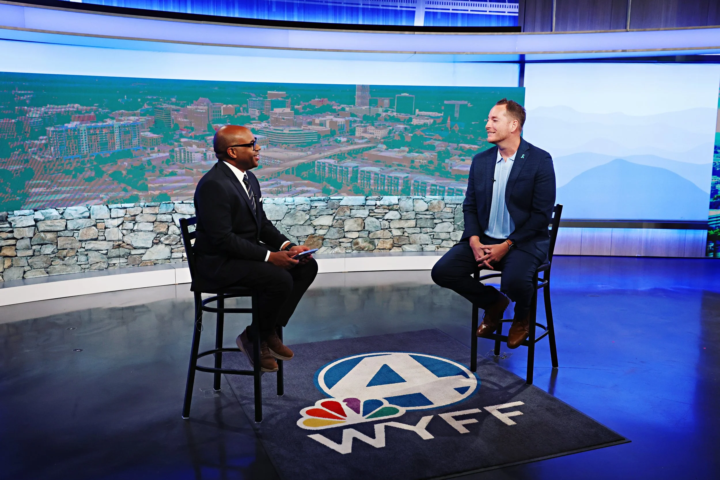 Two men seated and talking in a TV studio with a large background image of a cityscape and a NBC logo and text on the floor.