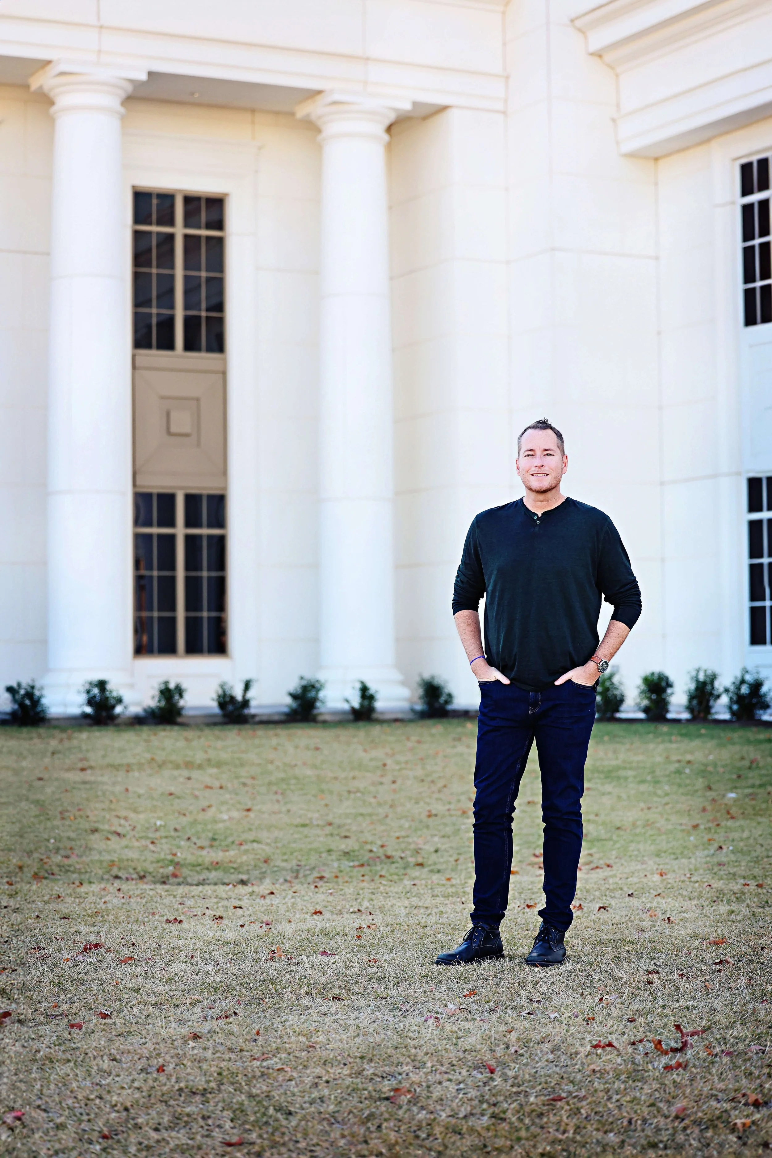 A man standing on a lawn in front of a large white building with tall columns and rectangular windows.